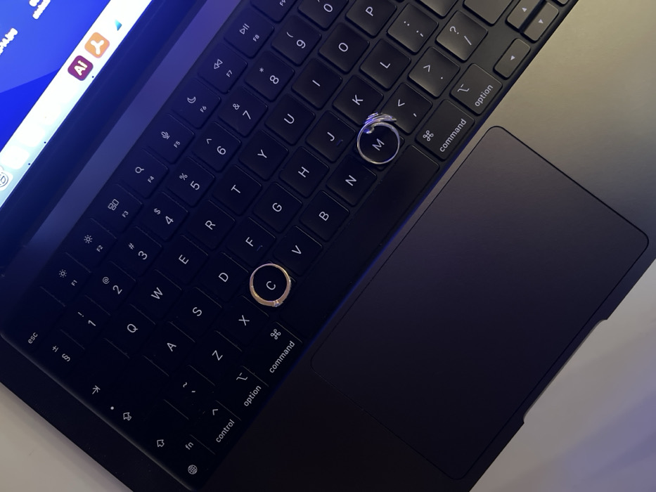 The scene depicts a dark-colored laptop, possibly a MacBook, positioned on a light-colored surface in the city of Bar, Montenegro. The laptop's dark QWERTY keyboard is prominently visible, angled from an overhead perspective. Two rings are resting on the keyboard: a gold-toned ring with a small, clear stone is centered on the 'C' key, and a silver-toned ring is centered on the 'M' key. A portion of the laptop screen is visible at the top, displaying a blue background with several application icons, including those resembling Adobe Illustrator and Adobe Acrobat Reader. The screen emits a distinct blue light that illuminates parts of the keyboard and the immediate surroundings. The laptop's trackpad is visible in the lower-left quadrant, consistent with its dark color. No individuals or active interactions are observable.