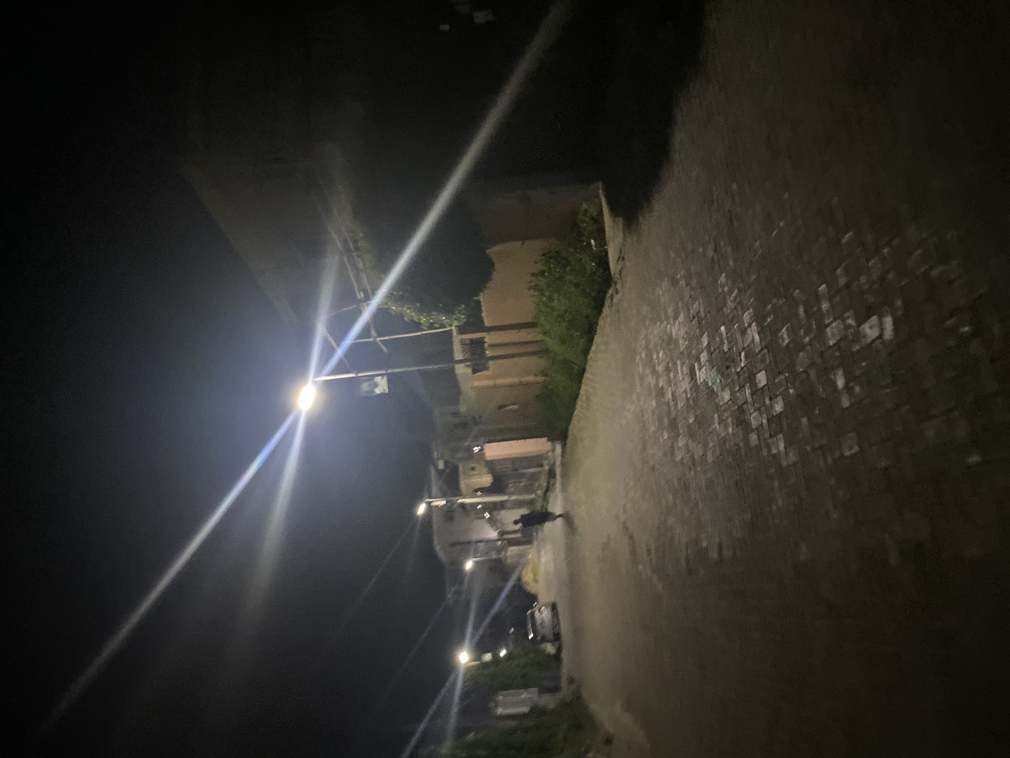 The image depicts a dimly lit street in Unknown, Unknown, at night. The street is paved with bricks and appears to have a sidewalk along the right side. Streetlights cast a bright glow, illuminating the path ahead. Several buildings can be seen in the background, with trees and bushes growing near them. A person is walking along the street, and a car is parked nearby. The overall environment seems quiet and residential.