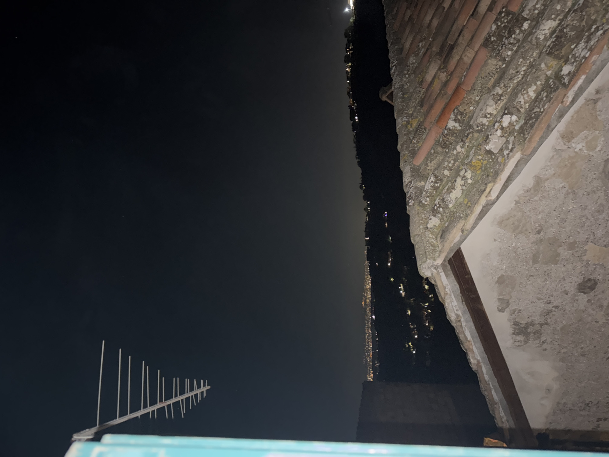 View from a rooftop in Castelnuovo di Porto, Italy at night. The night sky is dark with a few visible stars, an antenna is mounted on a blue-painted structure at the bottom of the frame. In the distance, the city lights are visible in a hazy, orange glow. The rooftop edge is on the right, showing aged bricks and weathered concrete. The bricks are red and covered with lichen and moss. The concrete is stained.