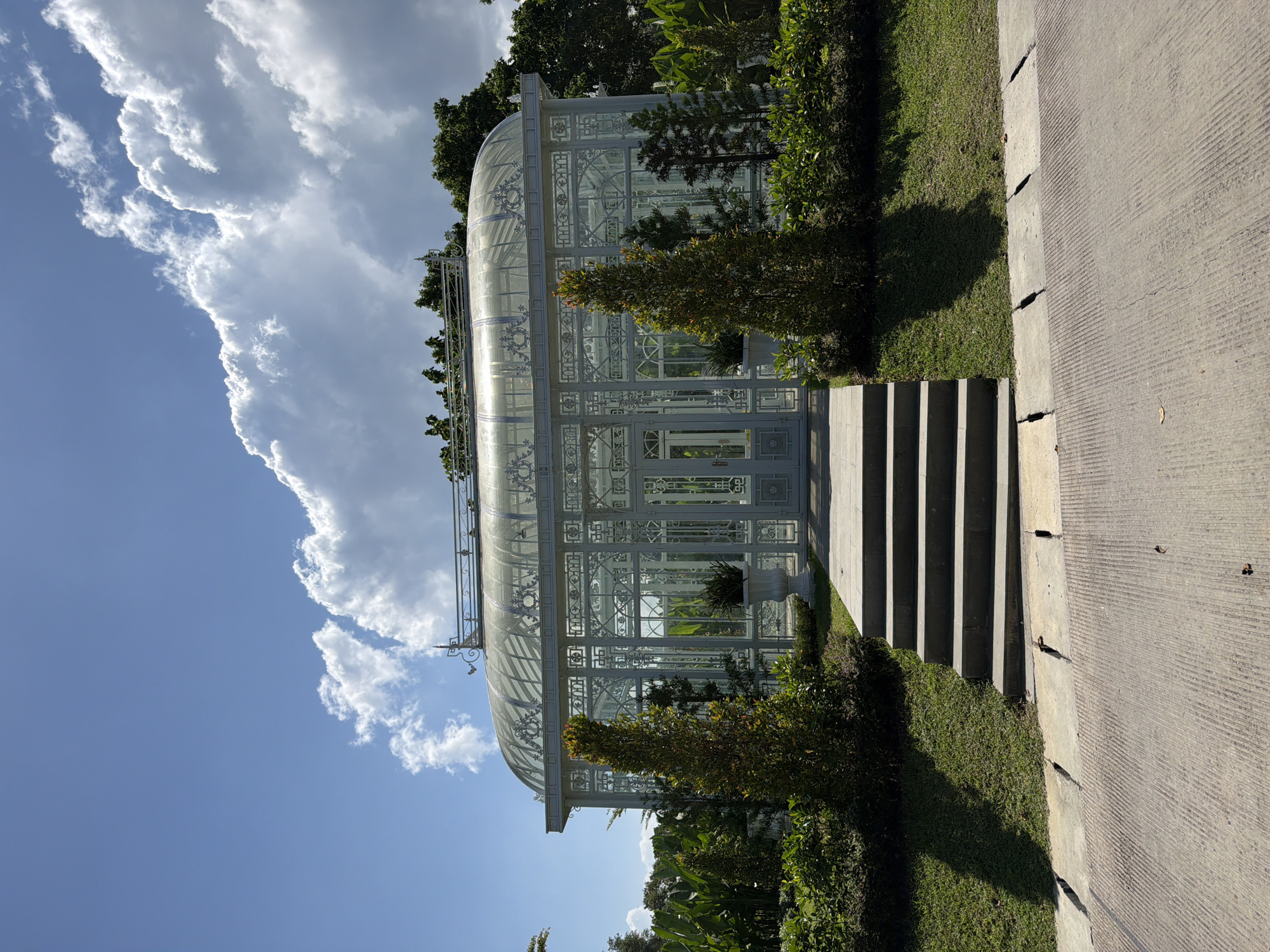 In Kediri, Indonesia, a large, ornate white greenhouse stands under a bright blue sky with fluffy white clouds. The greenhouse features a curved glass roof and intricate white metal framing, with double doors at the entrance. In front of the greenhouse, a set of stone steps leads up from a paved pathway to a garden area filled with lush greenery and two conical trees. The scene is bathed in sunlight, casting shadows on the grass.
