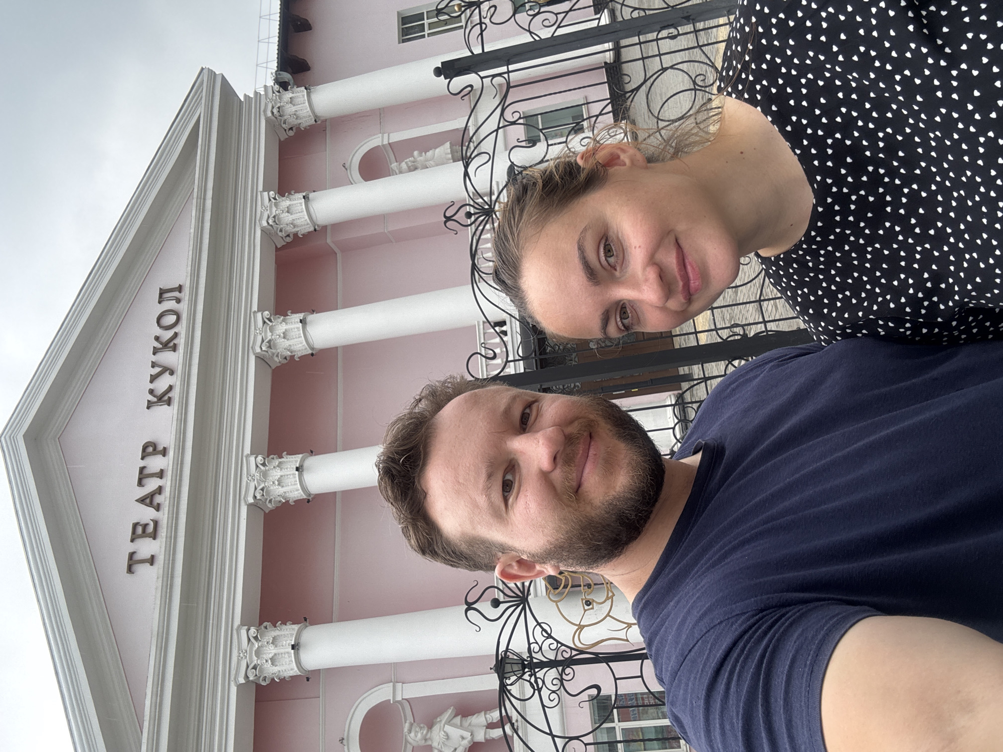 A selfie is taken by a man and a woman in front of the Blagoveshchensk Puppet Theatre in Blagoveshchensk, Russia. The man, on the left, wears a dark blue t-shirt and has a beard. The woman, on the right, has blonde hair and wears a black and white polka dot top. Behind them, the pink building of the theatre is visible with white columns and decorative script above. A black metal fence with ornate scrollwork is in the foreground.
