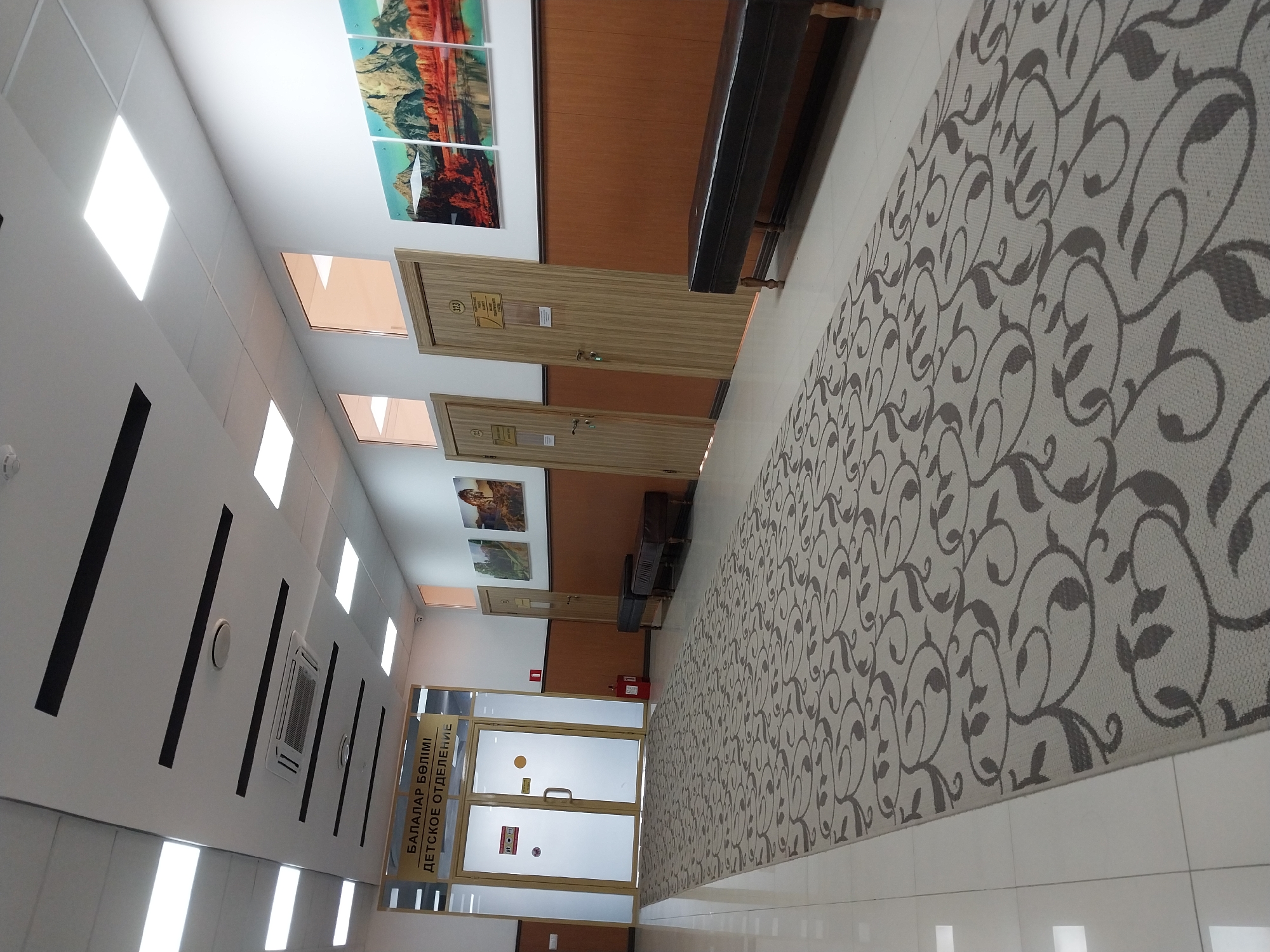 The image depicts an interior hallway of what appears to be a public building. The hallway is long and well-lit, with a patterned carpet running down the center. On the left side of the hallway, there is a set of glass double doors with a sign above them written in both Kazakh and Russian, indicating "Children's Department." There is a red fire extinguisher visible next to these doors. Further down the hallway, there are several closed wooden doors, some with numbers and signs on them. On the right side of the hallway, there are three framed landscape images mounted on the wall, depicting mountains and water. A long, dark brown bench is situated against the wall. The walls are a light color, with a darker brown wainscoting in some areas. The ceiling is white with recessed lighting panels and black linear accents, and an air conditioning unit is visible. The floor is tiled with white square tiles. No people are visible in the image. The presence of signage in Kazakh and Russian suggests the location is in Kazakhstan, and specifically in the city of Oral, given the linguistic context.