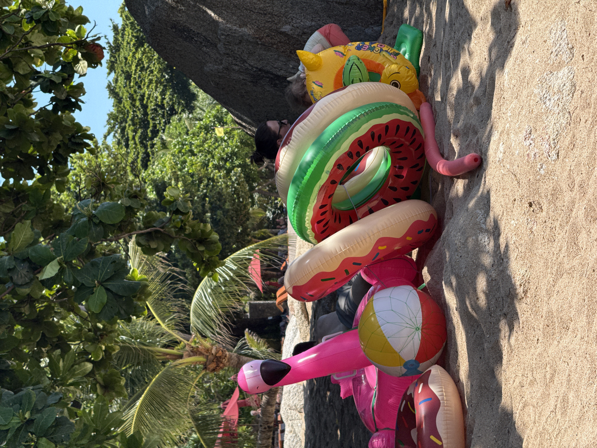 This image depicts a collection of colorful inflatable toys scattered on a sandy beach in the Ko Samui District, Thailand, likely near Bophut or Chaweng Beach, areas known for their resorts and tourist activities. A bright pink inflatable flamingo, a beach ball with yellow, white, and red panels, and several inflatable rings designed to resemble donuts and slices of watermelon are prominent in the foreground. Behind the toys, partially obscured by foliage and a large boulder, are two individuals, a woman wearing sunglasses and a child. In the background, palm trees and lush green vegetation suggest a tropical environment, with red umbrellas indicating a resort or beachside establishment. The scene is bathed in sunlight, casting shadows from the foliage onto the sand.