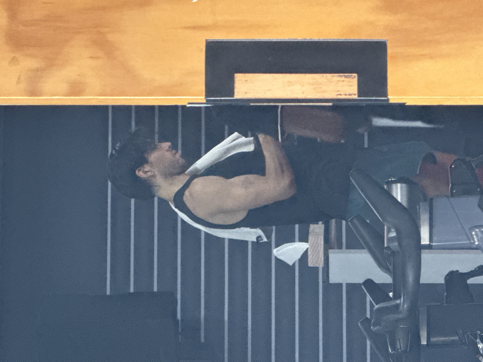 This image depicts a man exercising indoors, likely at a gym in Puebla, Mexico. The man is shown in profile, seated on an exercise bike and facing towards the left side of the frame. He has dark hair and visible stubble or a short beard. He is wearing a black tank top and dark shorts, with a white towel draped over his shoulders and neck. His right arm, prominently featured, displays muscle definition, suggesting active engagement in his workout.
Adjacent to the man, on the right, components of the exercise bike are visible, including handlebars and a dark metal water bottle secured in a holder on the bike's frame. The background consists of a dark wall with notable light-colored vertical stripes, which commonly feature in gym aesthetics. Above this striped wall, there is a broad, light-brown wooden panel, possibly part of the ceiling or an upper wall section. A dark, rectangular object, potentially a monitor or a piece of equipment, is situated on another lighter wooden surface in the upper background. The lighting suggests a well-illuminated indoor setting. There is no visible text in the image. The man's posture indicates focus on his physical activity.