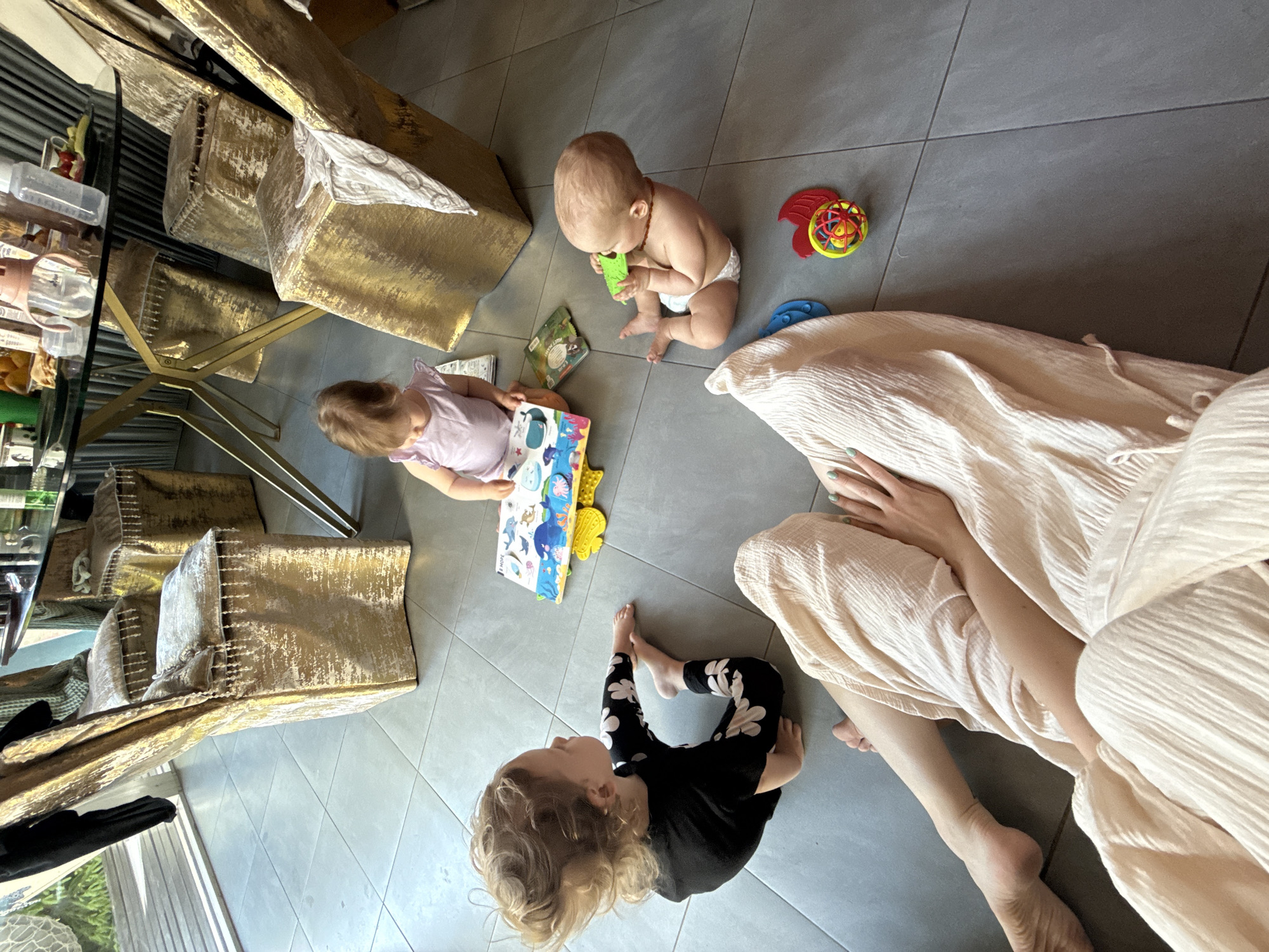 Observed indoors, the scene features three children on a gray tiled floor near gold cushioned furniture. A baby, wearing a diaper, is chewing on a green object. A toddler in a pink shirt is looking at a book, while another toddler with curly hair, dressed in black and white patterned leggings and a black shirt, looks upward. A woman is present, wearing a light-colored dress or robe, sitting on the floor with her legs visible. Toys are scattered around the children. The location is not Чёрное, Russia, as the objects in the image provide no direct link or visual evidence to this location.