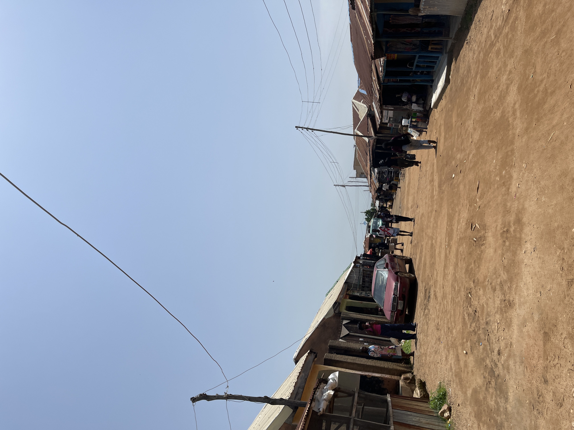 The image depicts a street scene in Jos, Nigeria, likely a market or commercial area. The ground is unpaved and dusty, with a reddish-brown color. On either side of the street are numerous small shops or stalls with corrugated metal roofs and open fronts. Some structures appear to be constructed from wood and metal.
Several people are visible walking along the street and standing in front of the shops. A red sedan is parked on the left side of the street, partially obscured by a structure. Overhead, power lines are strung across poles, indicating the presence of electricity. The sky is clear and bright, suggesting a sunny day. The overall impression is of a bustling, informal commercial environment.