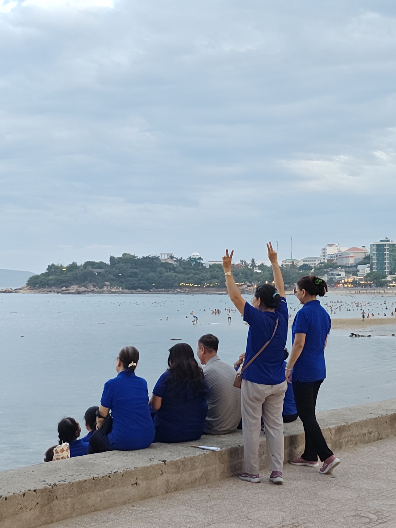 A group of people are sitting and standing on a concrete walkway overlooking a crowded beach and the sea in Nha Trang, Vietnam. In the foreground, two women in blue shirts are standing. One woman on the left is raising both hands with two fingers extended on each hand in a peace sign. The other woman on the right is looking to her left. Behind them, several people are seated on the walkway, including a woman in a blue shirt facing away from the camera, a man in a gray striped shirt, and two children. The beach in the background is filled with numerous people, and buildings and palm trees are visible on the coastline. The sky is overcast with clouds.