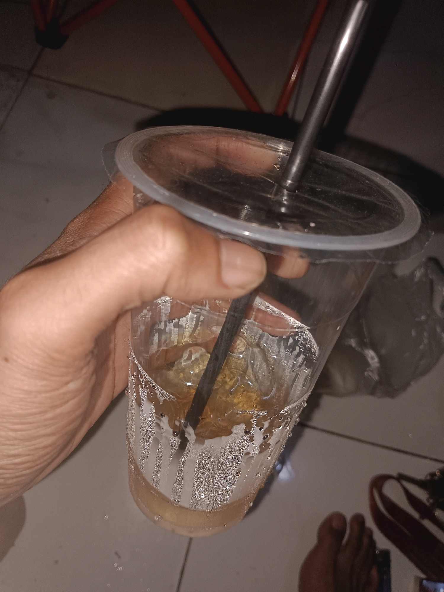 A close-up, indoor shot shows a person's hand holding a clear plastic cup filled with a yellow-amber colored liquid and ice. A metal straw is inserted into the drink. The cup is sweating, with condensation visible on the outside. The background is dimly lit and appears to be a tiled floor. Part of a red chair is visible in the upper left corner. The overall lighting is dim, suggesting it might be evening or a poorly lit indoor space. The location is Palimanan, Indonesia.