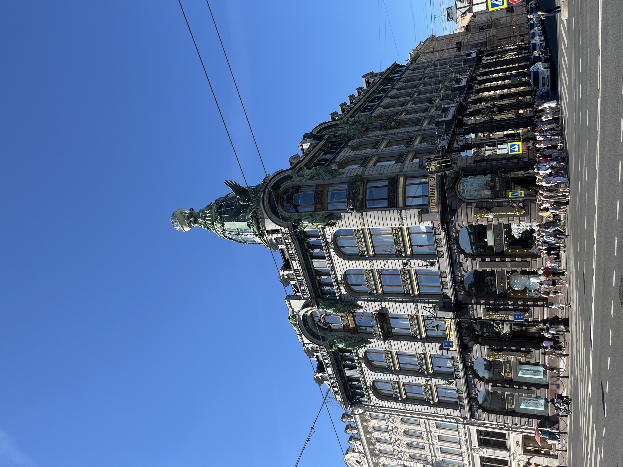 This is a low-angle, wide shot of the Singer House, also known as the Dom Knigi, located on Nevsky Prospekt in Saint Petersburg, Russia. The grand, ornate building features classical architecture with art nouveau elements, characterized by its imposing facade, numerous windows, and a distinctive glass dome topped with a globe and an eagle sculpture. The building is situated at a busy intersection where people are walking on the sidewalks and crossing the street. Vehicles, including cars and a scooter, are visible on the road. The sky is clear and blue. The building's exterior is adorned with sculptures of figures and decorative elements. Businesses such as "MK store" and "Cafe René" are identifiable on the ground floor. The scene captures a bustling urban environment under bright daylight.