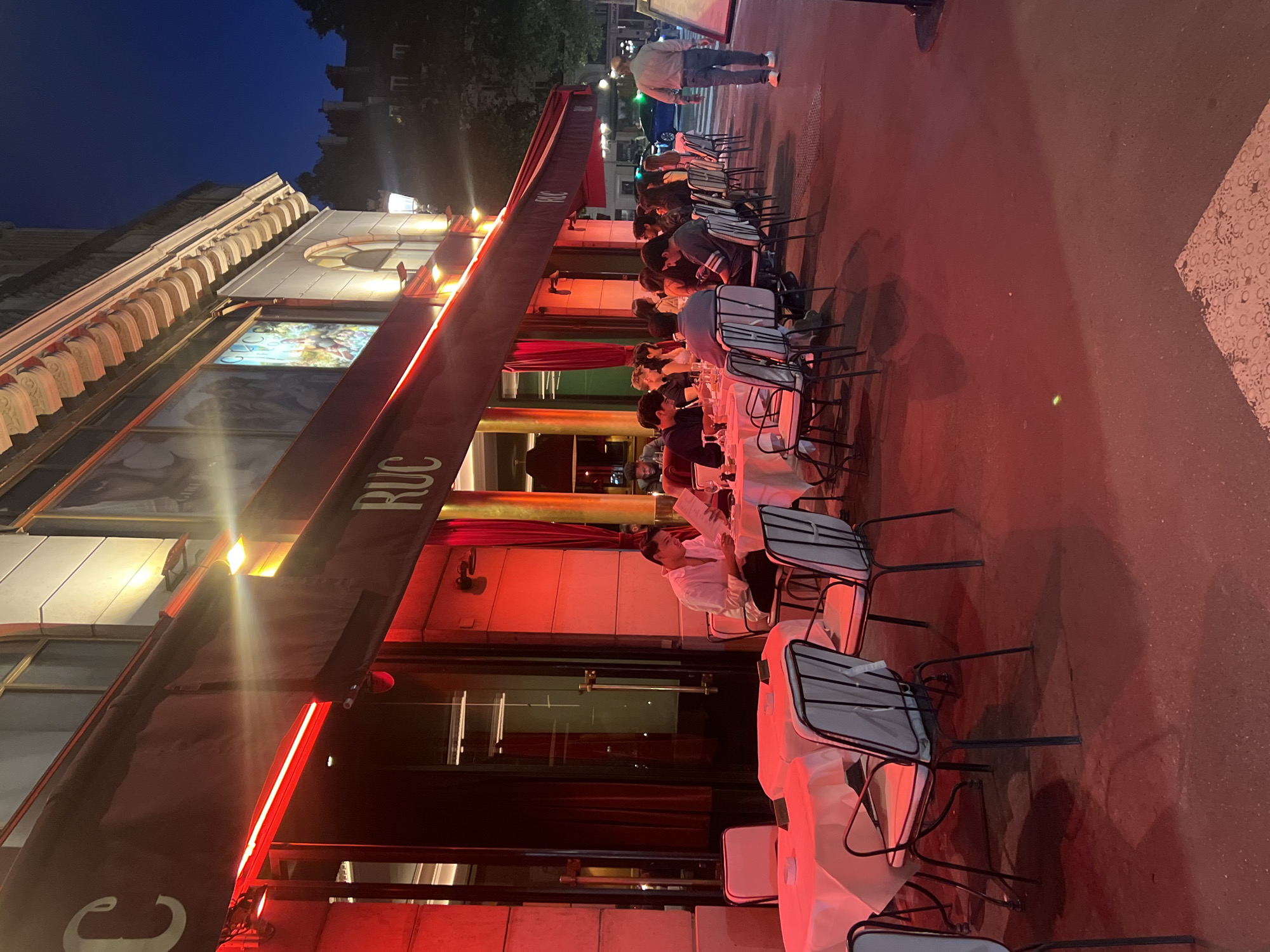 The image depicts an outdoor seating area of a restaurant at night in Paris, France. Red neon lights illuminate the scene, casting a vibrant glow on the facade of the building and the tables. The restaurant appears to be situated on a street, with a few people dining at tables covered with white tablecloths. The building facade features large windows and ornate architectural details. In the background, a few trees and other buildings are visible, suggesting an urban setting. A person is seen walking away from the seating area on the sidewalk. The overall ambiance is lively and inviting, characteristic of Parisian nightlife.