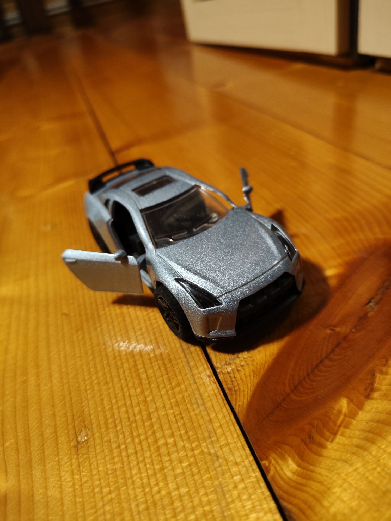 A close-up, high-angle shot shows a silver toy car model with its driver's side door ajar, resting on a polished wooden surface. The car is angled diagonally across the frame, with its front facing toward the bottom right. The wooden surface exhibits a prominent wood grain pattern with varying shades of orange and yellow, suggesting it might be a table or floor. Subtle reflections and light glint off the polished wood. The background is blurred, with hints of a lighter colored object and a darker surface. There are no people or animals present in the image. The scene appears to be indoors, possibly in a home or workshop, given the wooden surface and the presence of a toy. The location in Фестивальное Снт, Russia, is not directly evident from the visual information within the image itself.