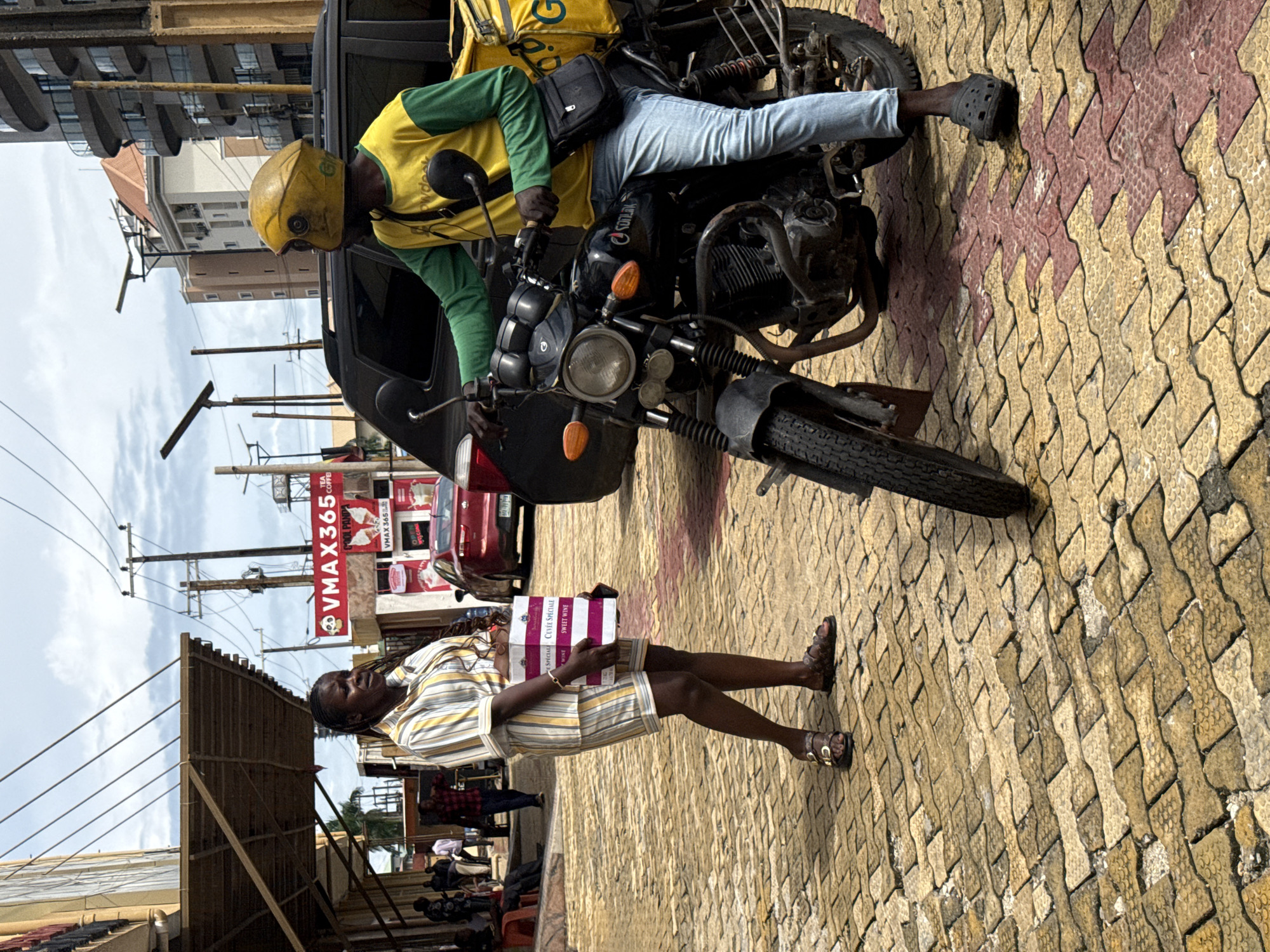 In Itirin, Nigeria, a woman in a striped yellow and white shorts set stands on a textured yellow and red brick sidewalk, holding two stacked white boxes with pink lettering that reads "CUVEE SPECIALE SWEET WINE." To her right, a delivery person wearing a yellow and green long-sleeved shirt, light blue jeans, and a yellow helmet sits on a black motorcycle with a yellow delivery bag strapped to the back. The motorcycle has orange turn signals and the name "SONLAT" visible on the side. Behind them, a black SUV is parked. The background shows a cloudy sky, power lines, buildings, and a sign that reads "VMAX365 TEA COFFEE." Other people are visible in the distance, some near a red car. The scene appears to be an outdoor market or street.