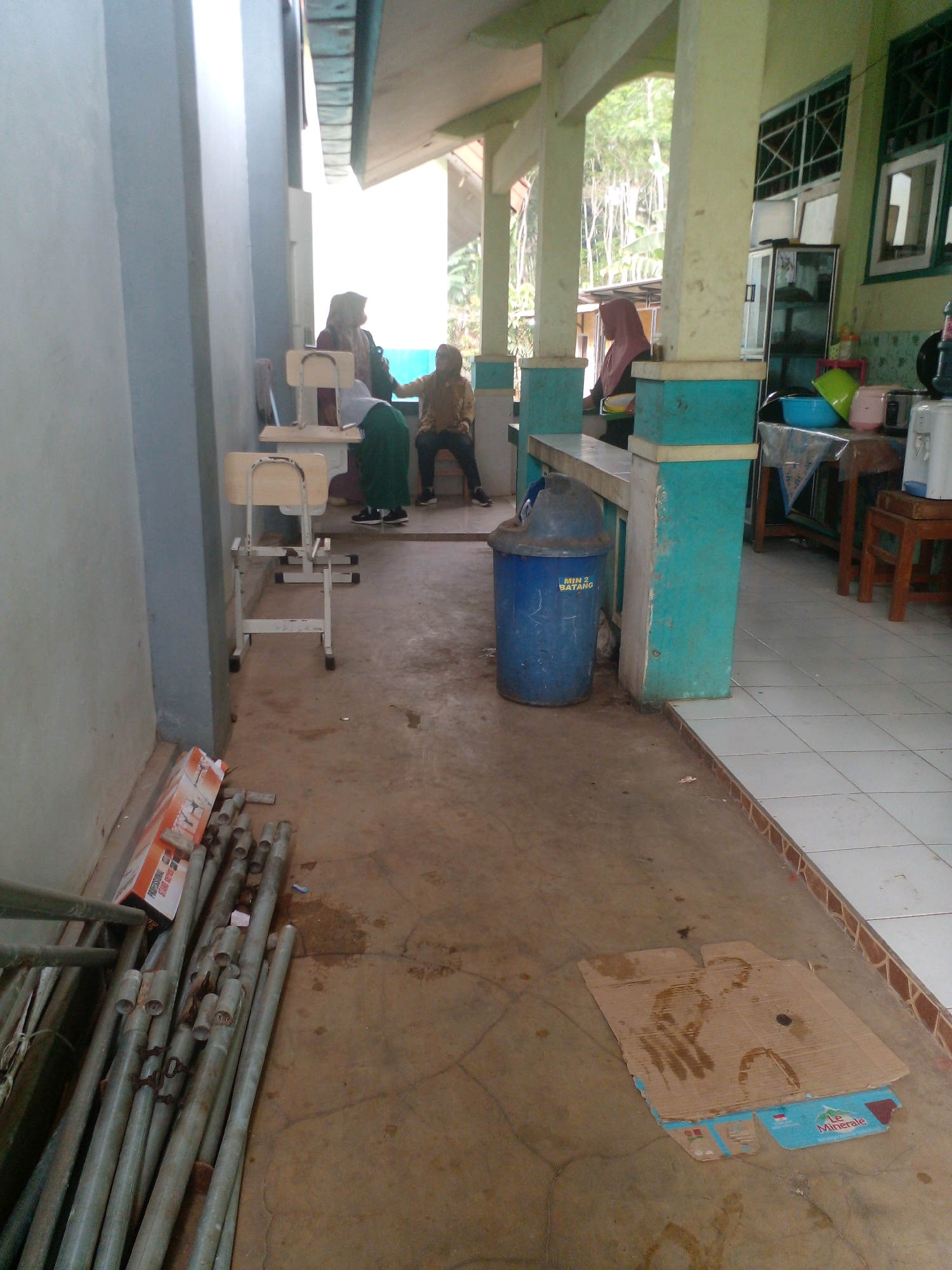 The image shows an outdoor area that appears to be a school hallway or covered walkway in Banyuputih, Indonesia. In the background, three individuals are present. One person is seated on a chair and wearing a green garment. Another person is seated on a bench, wearing a light brown long-sleeved shirt and dark pants. The third individual is standing. A blue trash can with "MIN 2 BATANG" written on it is visible on the right side of the frame. Several metal pipes and a cardboard box are lying on the ground in the foreground. The walkway has a concrete floor and pillars with turquoise accents. The environment suggests a casual setting, possibly during a school day.