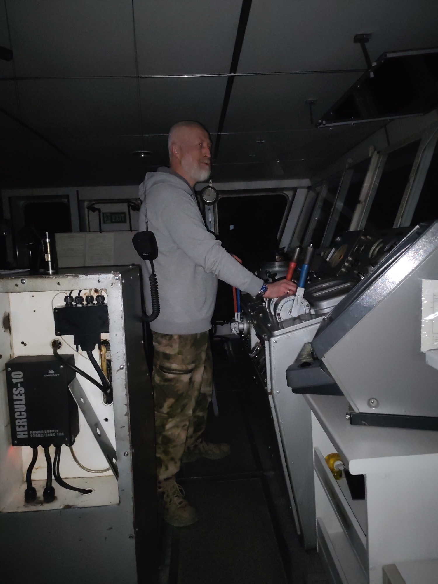 This image, taken in Golyashi, Russia, shows a man standing at the controls of what appears to be a ship's bridge or control room. The setting is dimly lit, suggesting nighttime or a dark environment.
The main subject is a middle-aged man with a shaved head and a white beard, wearing a light gray hooded sweatshirt and camouflage-patterned trousers. He stands facing right, with his right hand resting on a control panel that features several levers, some with red and blue accents. A radio microphone with a coiled cord is clipped to his hip.
To the left of the frame, there is an open console or cabinet containing electronic equipment. A black box is visible with the text "HERCULES-10" and "UNICONT", indicating it's a "POWER SUPPLY 220VAC/24VDC 10A". Cables are connected to this device. An "EXIT" sign can be seen further back above a doorway. Above the main control area and windows, a dark, rectangular screen or monitor is mounted on the ceiling. The windows of the bridge are visible, looking out into darkness.
The man appears to be actively operating the controls of the vessel or machinery, his gaze directed forward, presumably out into the dark. The overall atmosphere is operational and focused, characteristic of a maritime or industrial setting during low visibility.
