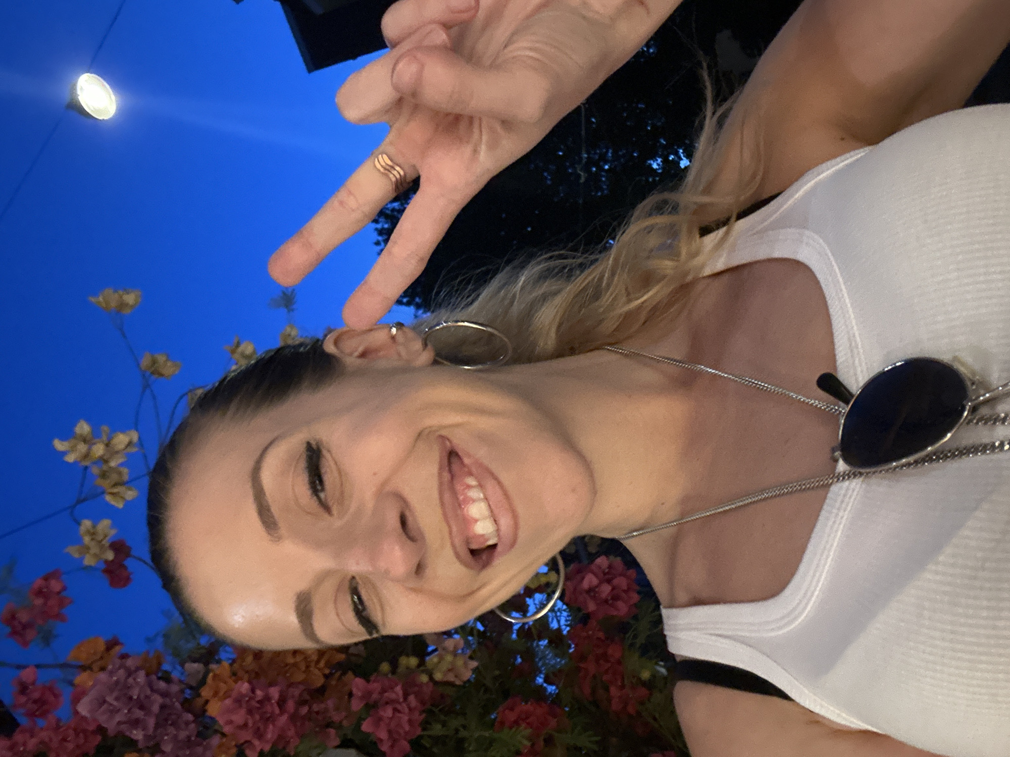 In a selfie taken at dusk, a woman with blonde hair pulled back in a ponytail smiles with her mouth open, revealing her teeth. She is wearing a white ribbed tank top and a silver necklace with sunglasses hanging from it. Her right hand is held up, forming a peace sign, with a wavy ring on her middle finger. She is adorned with large hoop earrings. Behind her, a vibrant display of pink and red flowers cascades, set against a deep blue evening sky. A single light bulb hangs in the distance, illuminating the scene. The overall impression is that of a candid moment captured outdoors, possibly in a lively urban setting like Amsterdam, Netherlands, given the presence of flowers and street lighting.