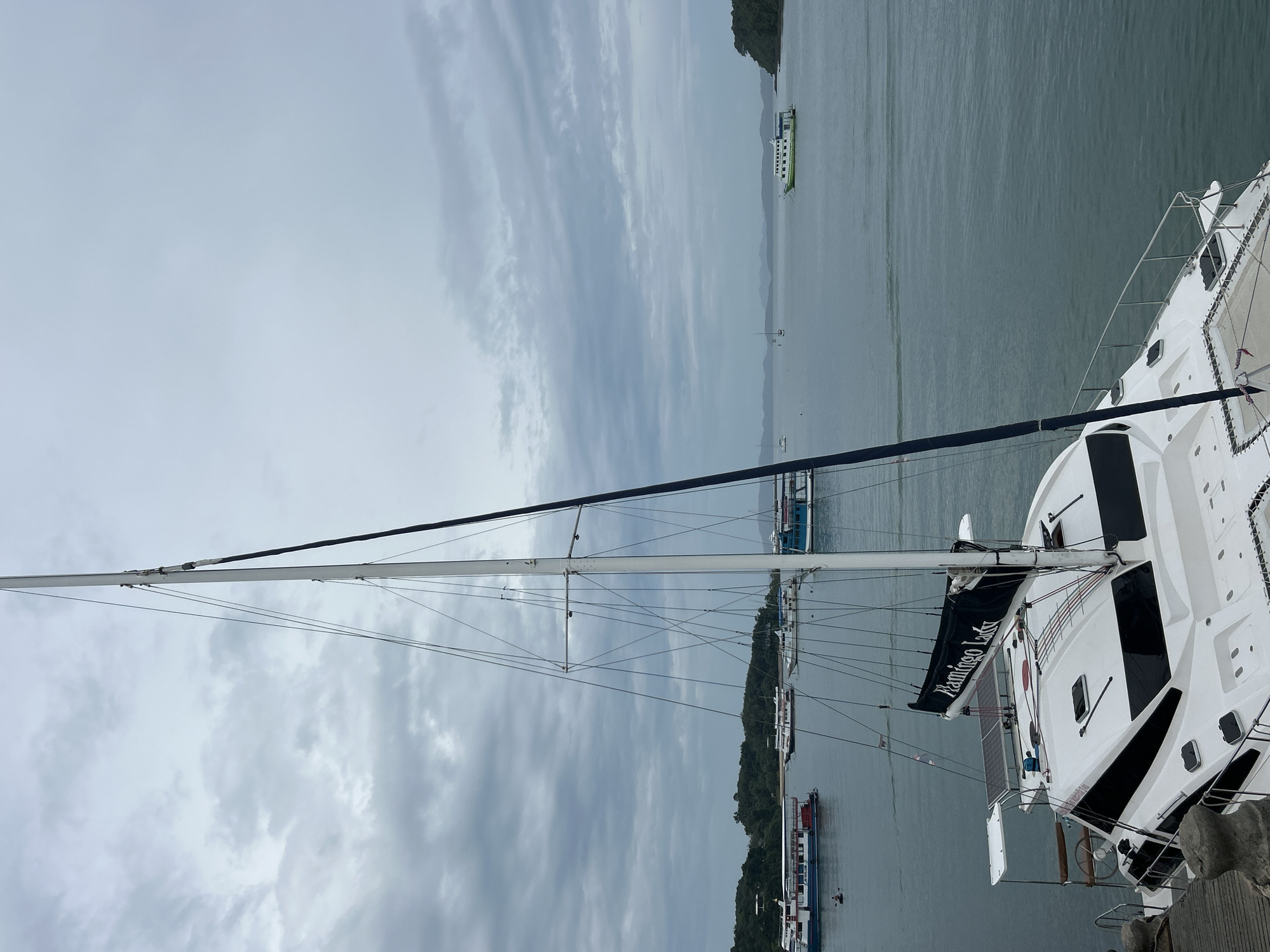 This image, taken in Pa Khlok, Thailand, depicts a cloudy day at a harbor filled with various boats. In the foreground, a white catamaran sailboat named "Flamingo Lady" is docked. The catamaran has a tall mast and dark-colored sails, one of which displays the boat's name in white lettering. The deck of the catamaran features solar panels and various hatches and windows. In the middle ground, several other boats, including passenger ferries and smaller vessels, are anchored or moving in the water. In the background, a tree-covered island is visible on the left, and rolling hills can be seen in the distance on the right. The sea is calm and the sky is overcast. A single person is visible in a kayak near the passenger ferry on the left.