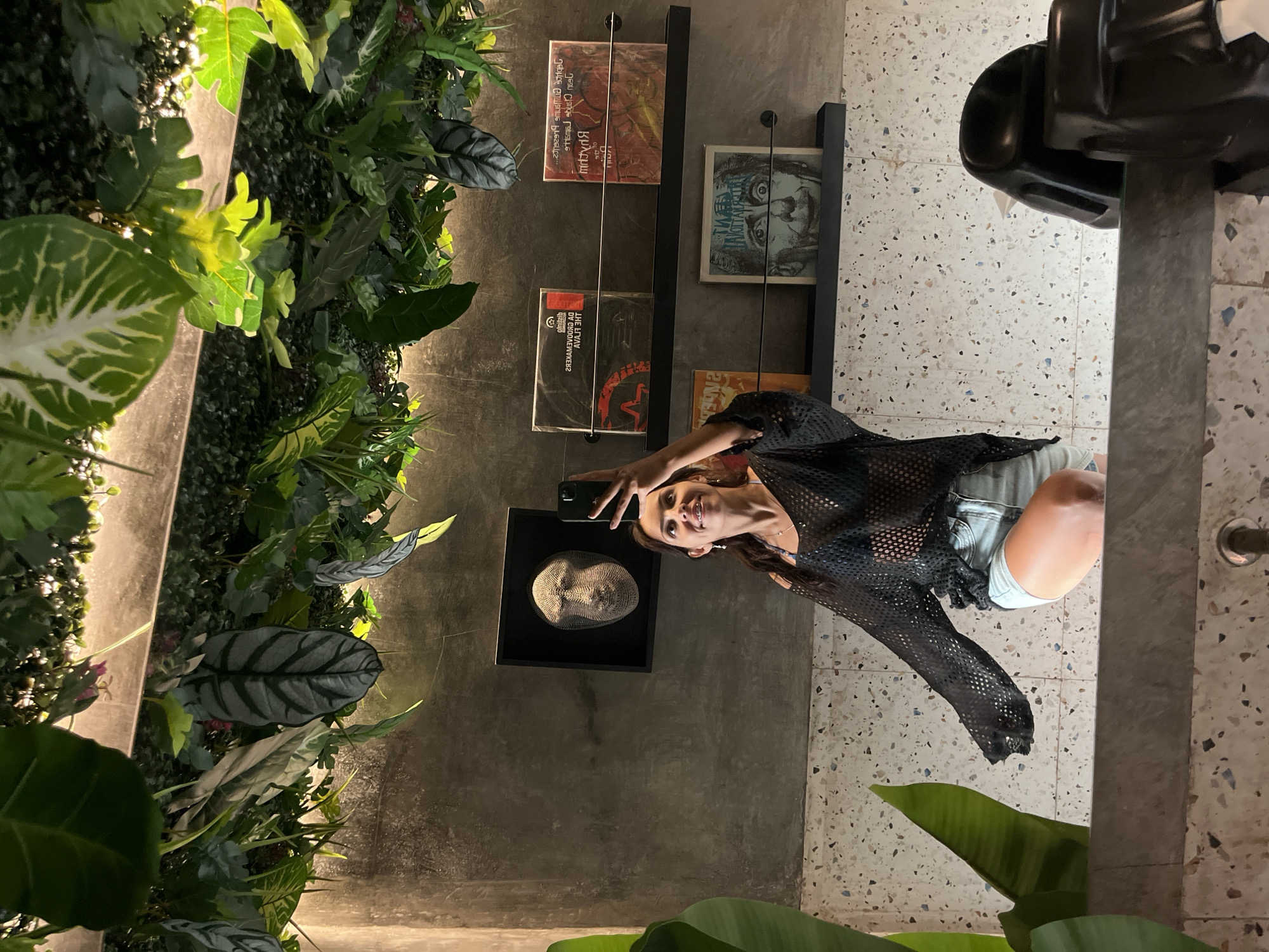 The image, when properly oriented, captures a young woman posing for a selfie in a chic indoor environment. She stands towards the right side of the frame, holding a smartphone above her head, looking up with a wide smile at the camera. She is dressed in a black mesh long-sleeved top layered over what appears to be a dark bikini top, paired with light-colored shorts.
The background is dominated by a dark grey wall. To the left, a vibrant vertical garden of artificial or real green plants is mounted, featuring various leaf shapes and textures, subtly lit from below by warm accent lights. On the right side of the wall, a black shelving unit holds several items, likely books or record covers, displayed vertically. Visible text includes Russian words "ВОЛЧОНОК ЛУИС" (meaning "Wolf Cub Luis") on one cover, and "AVALA" along with a mirrored "ЗНТ" (ZHT) on another. Below the shelves, a framed 3D sculpture of a human face is mounted on the wall. The floor is made of light-colored, speckled terrazzo tiles. A large black object, possibly a piece of furniture, is partially visible in the upper right corner. The overall lighting is subdued, with focused illumination enhancing the decorative elements of the space, suggesting a modern and artistic setting, possibly a cafe, bar, or lounge in Kuta Selatan, Indonesia. The woman projects a joyful and relaxed demeanor.