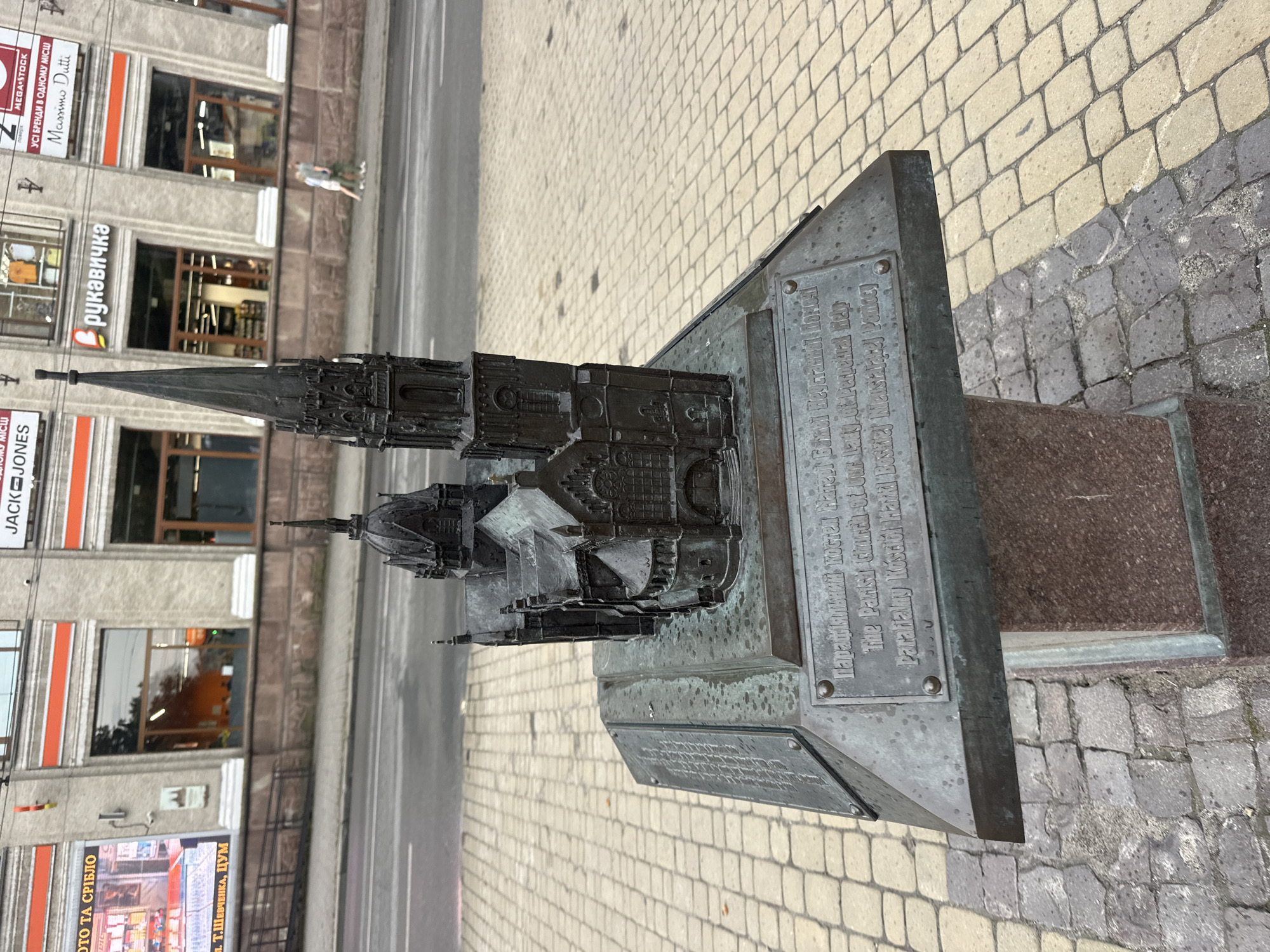 A close-up, eye-level shot features a detailed metal miniature of a church with a tall spire, placed on a textured metal base. The base itself rests on a granite pedestal. The church model is dark, weathered metal, with intricate architectural details. The base has two plaques; the top one is filled with text that is difficult to read, while the bottom plaque is clearer, displaying "The Parish Church of Our Lady of Perpetual Help" and "Parafiiălʹnyy̆ Kôstëlìlʹ Năshĭ Leydĭ Pomĭchnotsi." The surrounding ground is paved with interlocking beige and gray stones. In the background, a street with light traffic and a building with large windows and orange trim are visible. The building has signage including "JACK & JONES" and "рукавичка". A couple is walking on the sidewalk in the background. The overall impression is one of an urban setting, possibly a tourist spot, with the miniature serving as a representation of a local landmark. The plaque suggests the location is Ternopil, Ukraine, due to the presence of Ukrainian text and the recognizable style of the building and architecture, consistent with the city's historical center.