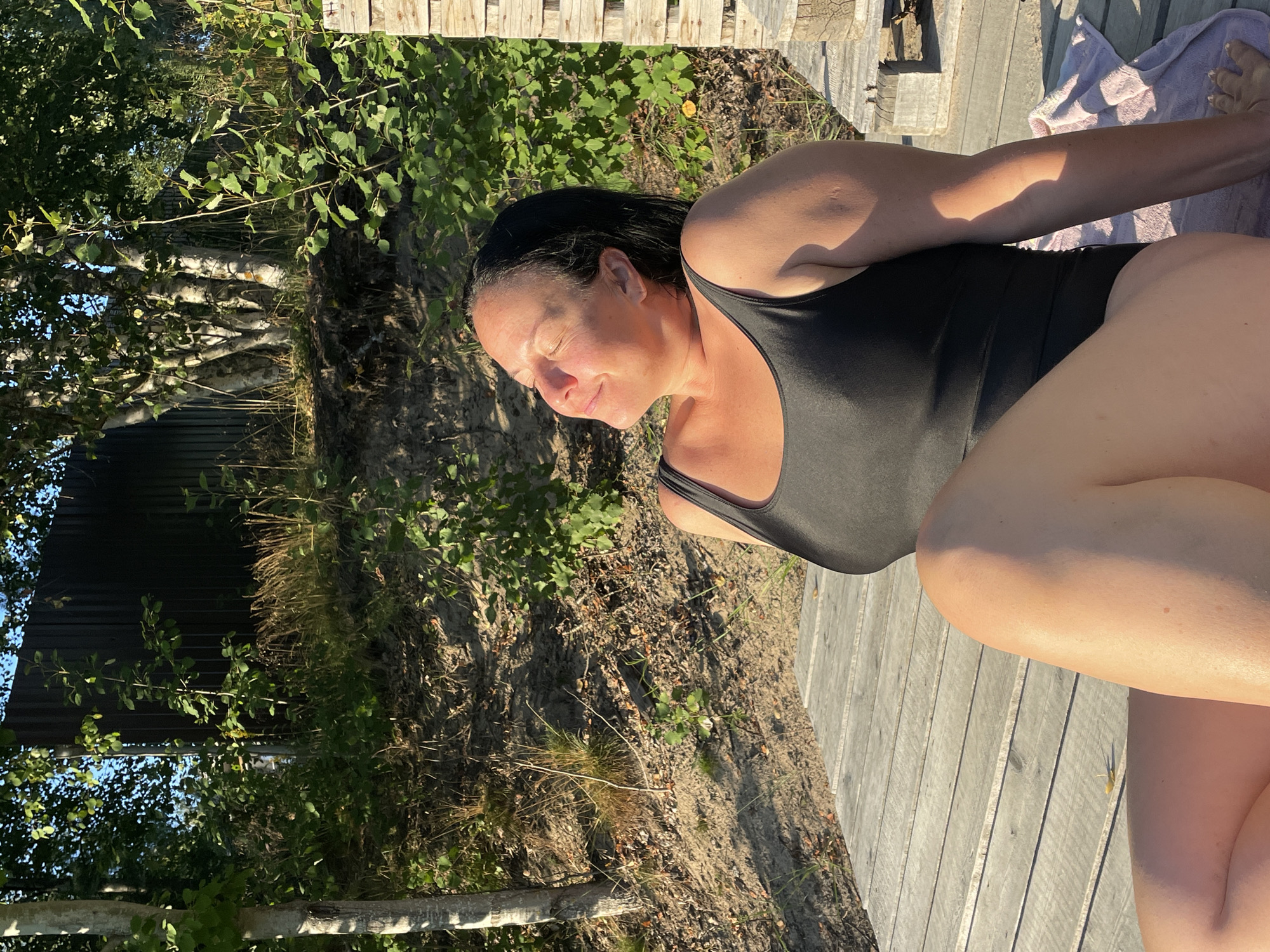 A woman sits on a wooden deck, facing the sun with her eyes closed. She is wearing a black one-piece swimsuit. The deck is made of weathered wooden planks. Behind her, there is a sandy slope covered with grass and trees. A dark metal fence is visible in the background, partially obscured by foliage. The scene appears to be outdoors, possibly near a body of water, given the sandy area and the presence of a towel on the deck. The lighting suggests it is daytime, with sunlight casting shadows. The location is identified as Kobona, Russia.