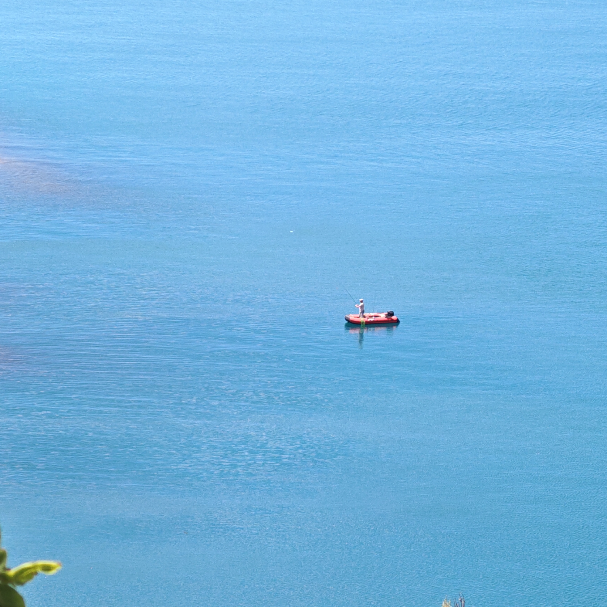 An aerial shot captures a bright, calm blue sea, reflecting a clear sky with slight ripples on the surface. In the middle ground, a small, red inflatable boat is occupied by a single person. The individual, dressed in light-colored clothing, stands in the boat, holding a fishing rod and appears to be actively fishing. An outboard motor is attached to the stern of the boat. To the far left, the blurry green foliage of a plant is visible, suggesting the viewpoint is from a higher elevation, possibly a cliff or hillside overlooking the sea. The overall scene is serene and evokes a sense of peaceful solitude. The location is the Black Sea near Sudak, Crimea, Ukraine.