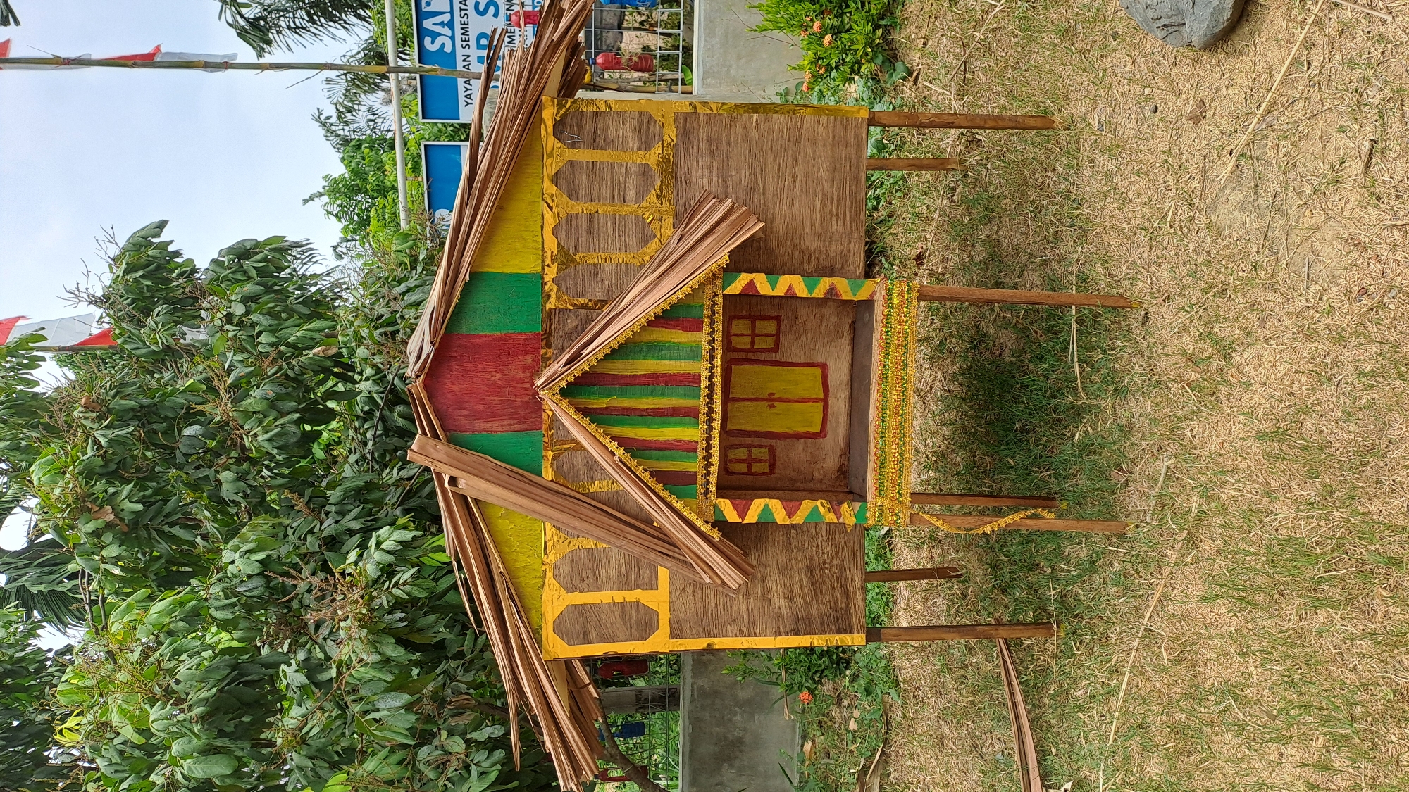 The image shows a miniature, stilt-raised house crafted from natural materials, likely displayed outdoors in Geulanggang Baro, Indonesia. The house features a layered roof thatched with dried palm leaves. The walls are constructed of wood-like material, possibly plywood, decorated with painted geometric patterns in yellow, red, and green. The front of the house has a smaller, more ornate entrance section with painted doors and windows. Decorative trim, possibly made of woven material, adorns the lower portion of the front facade and the raised platform. The structure is supported by four wooden stilts, planted in dry, patchy grass. In the background, lush green trees with some branches visible are present, along with a cloudy sky and glimpses of flags. Fencing and other structures are also faintly visible in the distance. The scene suggests a cultural display or craft project set in a semi-rural or outdoor community space.