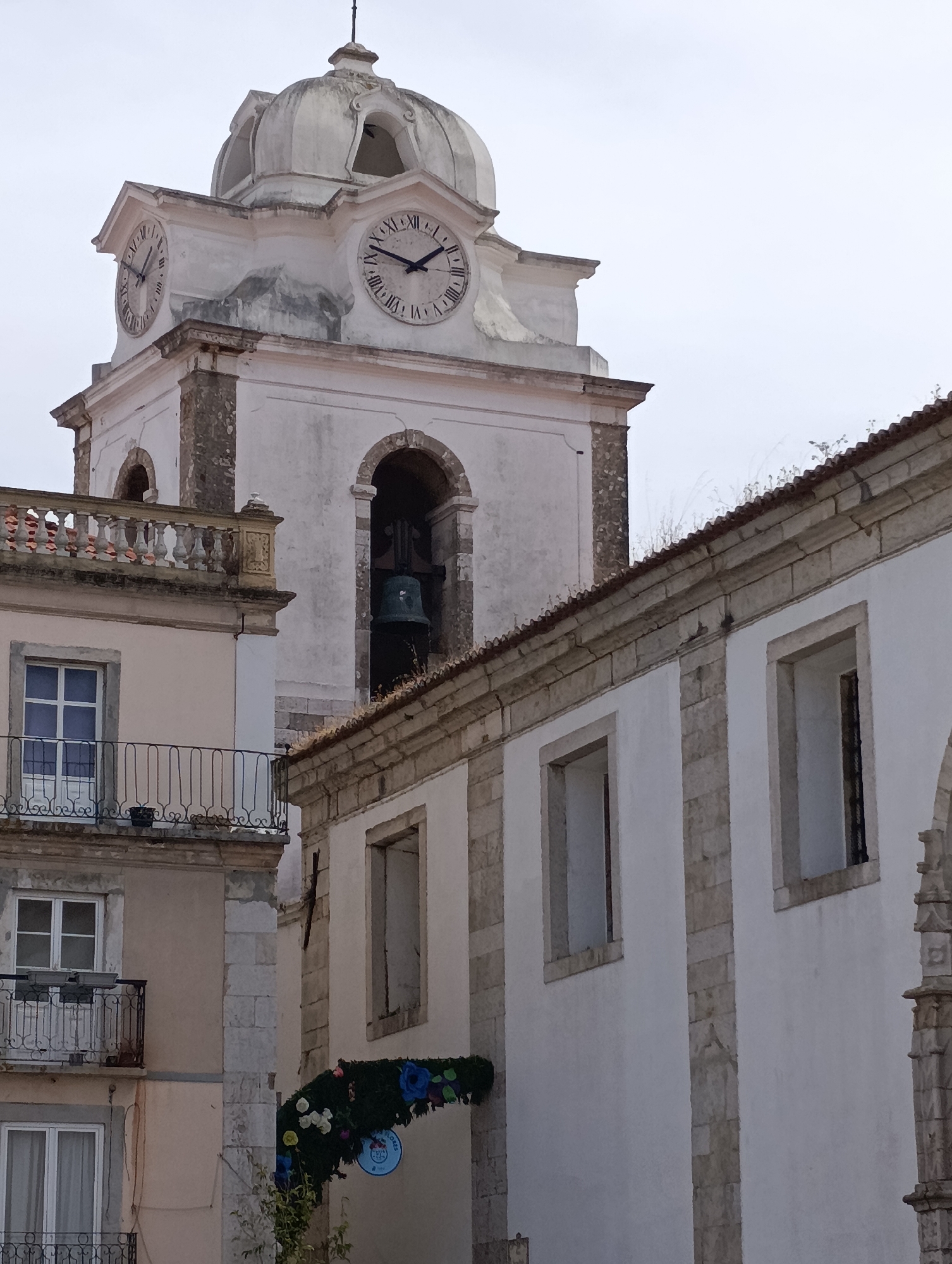 A bell tower of a white church with a domed roof stands tall against a pale sky. The tower has two large clock faces with Roman numerals; one shows 10:38 and the other 10:35. A large bell hangs in an arched opening within the tower. To the left of the bell tower is a light-colored building with a balcony. To the right is a white building with a textured roof and stone trim. A floral archway with blue and white flowers is visible in the lower center of the image, positioned in front of the white building. The overall scene is of an urban environment in Setúbal, Portugal, likely a historic district given the architecture of the church and surrounding buildings.