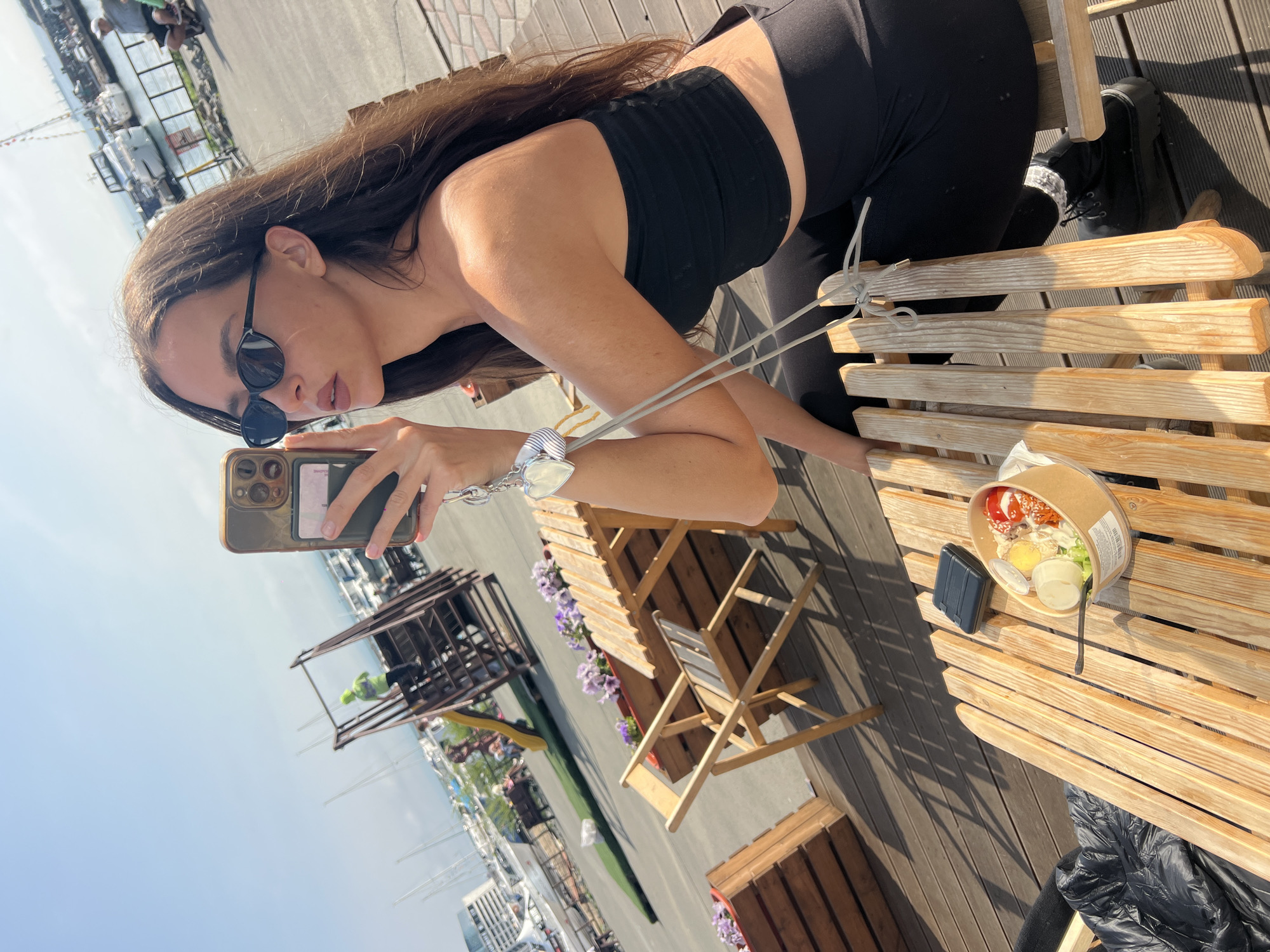 A young woman with long, dark hair is taking a selfie on a sunny day. She is wearing sunglasses and a black crop top. She is seated at a wooden table on a rooftop. On the table is a cardboard container with food and a black wallet. Below, there is a view of the harbor in Vladivostok, Russia, with numerous boats docked. There are flowers planted in wooden boxes near the seating area.