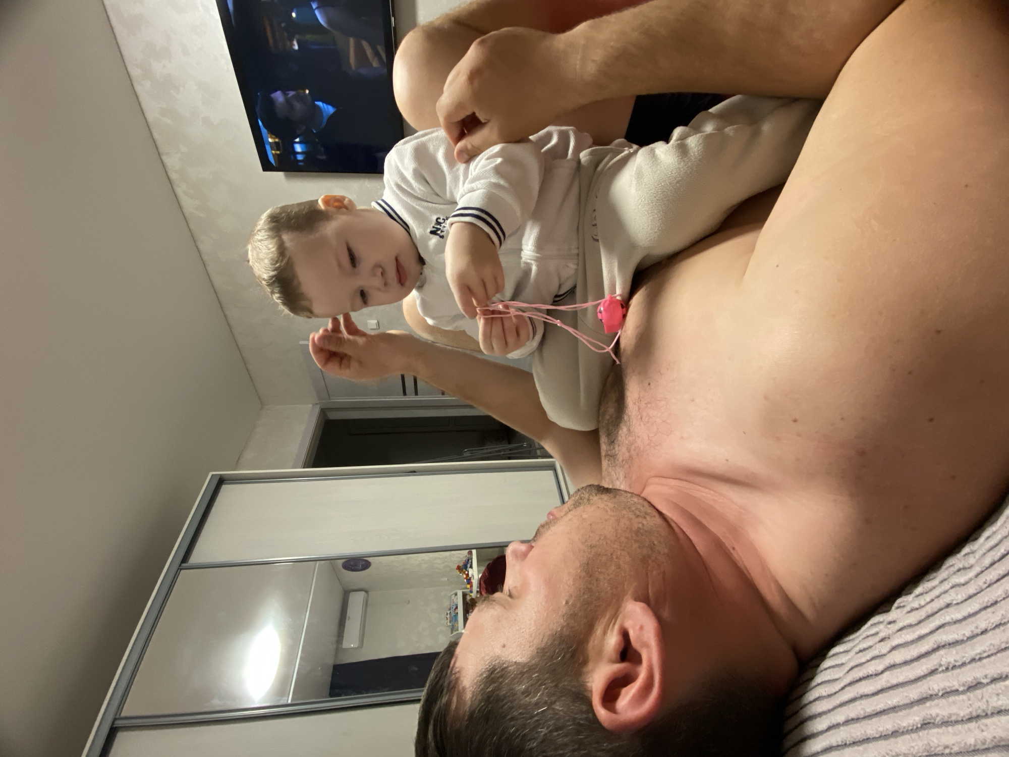 A man is lying on his back in a room. A young child is sitting on his chest, holding a pink object with strings. A television is visible in the background, and a mirrored sliding-door wardrobe is against the wall. The location is Sochi, Russia.