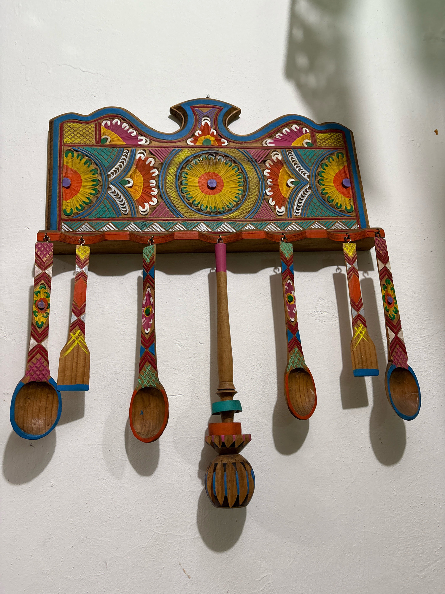 This is a photograph of a colorful, carved wooden decorative item depicting a set of spoons hanging from a decorative shelf. The item is mounted on a textured, off-white wall. The shelf itself is ornately decorated with a patterned design of stylized flowers and geometric shapes in vibrant colors, including blue, yellow, orange, red, green, and pink. Five wooden spoons hang from hooks on the underside of the shelf. Each spoon has a decorated handle with geometric patterns and bright colors, and the bowls of the spoons are unpainted wood, with some showing colored rims. The spoons vary slightly in size and shape. One spoon, hanging from the center, appears to be more decorative and elaborate than the others, with stacked, colored wooden elements. The lighting suggests the photograph was taken indoors, with a shadow cast on the wall behind the item, indicating a light source from the front and slightly to the right. This item is located in Cuernavaca, Mexico.