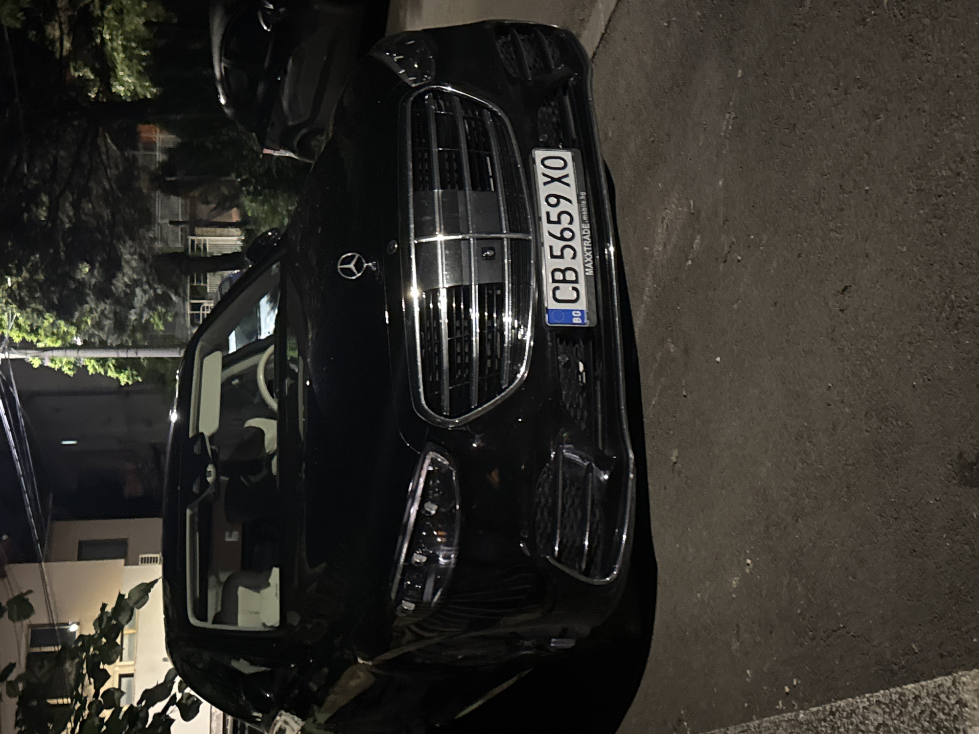 A black Mercedes-Benz is parked on the street, showing its front grill and license plate, which reads "CB 5659 XO" under the abbreviation "BG." The license plate also indicates a domain name, "MAXXTRADE mobile.bg." The car's interior features white seats and dashboard. The surroundings appear to be residential, with buildings and trees visible. The image was captured at night. The location is likely Sofia, Bulgaria, based on the "BG" abbreviation on the license plate.
