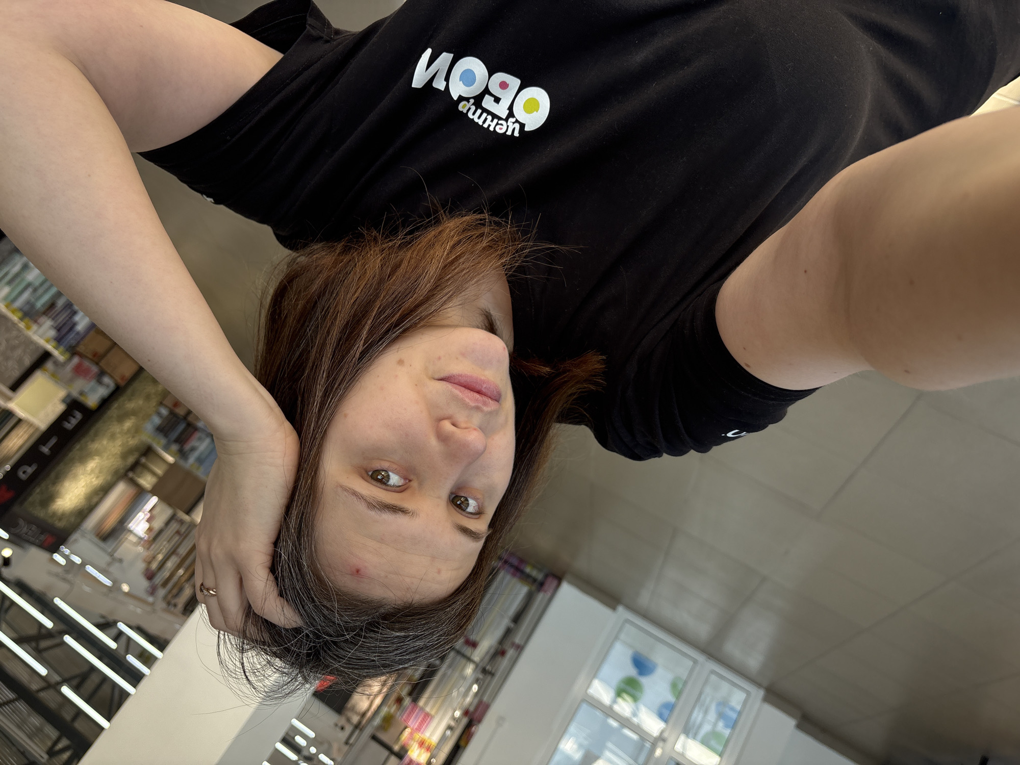 A woman is indoors, possibly in a commercial space in the city of Chaikovsky, Russia. She is wearing a black t-shirt with Cyrillic script and geometric shapes on it. Her dark hair has some grey strands. She is touching her hair with her hand that has a ring. She is looking at the camera. The background shows commercial shelves, items, and lighting, suggesting a retail setting.