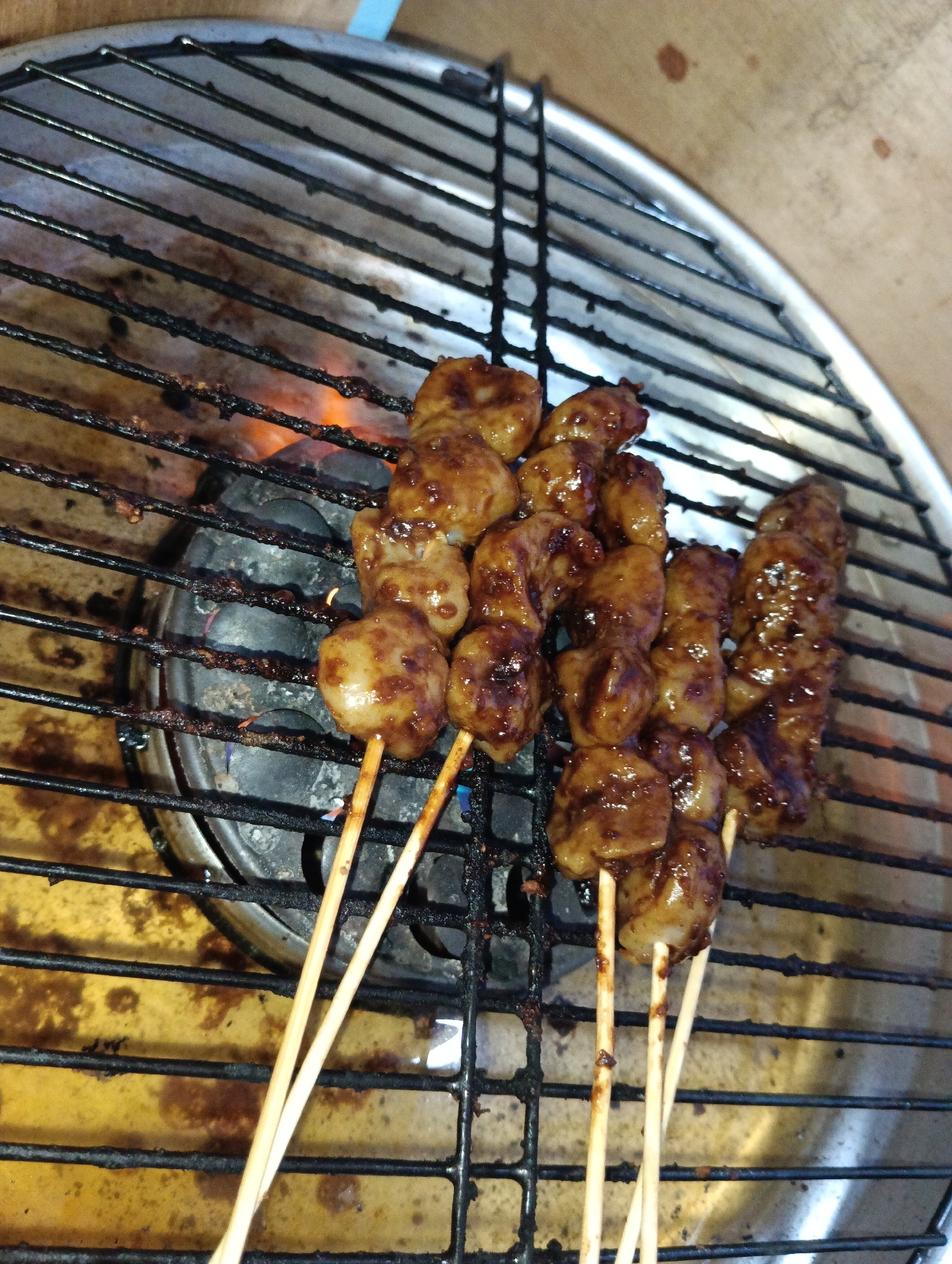 A close-up, overhead shot shows several skewers of grilled meatballs being cooked on a metal grill. The meatballs are small, round, and coated in a dark, glossy sauce. They are skewered on wooden sticks, and some of the sticks are charred from the heat. The grill grates are black and appear to be covered in grease and food residue. Below the grill, an orange flame is visible, indicating a gas burner or charcoal. The background is a light brown, wooden surface. There are no people visible in the image. The scene appears to be an outdoor food stall or a home barbecue. The location is in the city of Pematang Jaya, Indonesia.