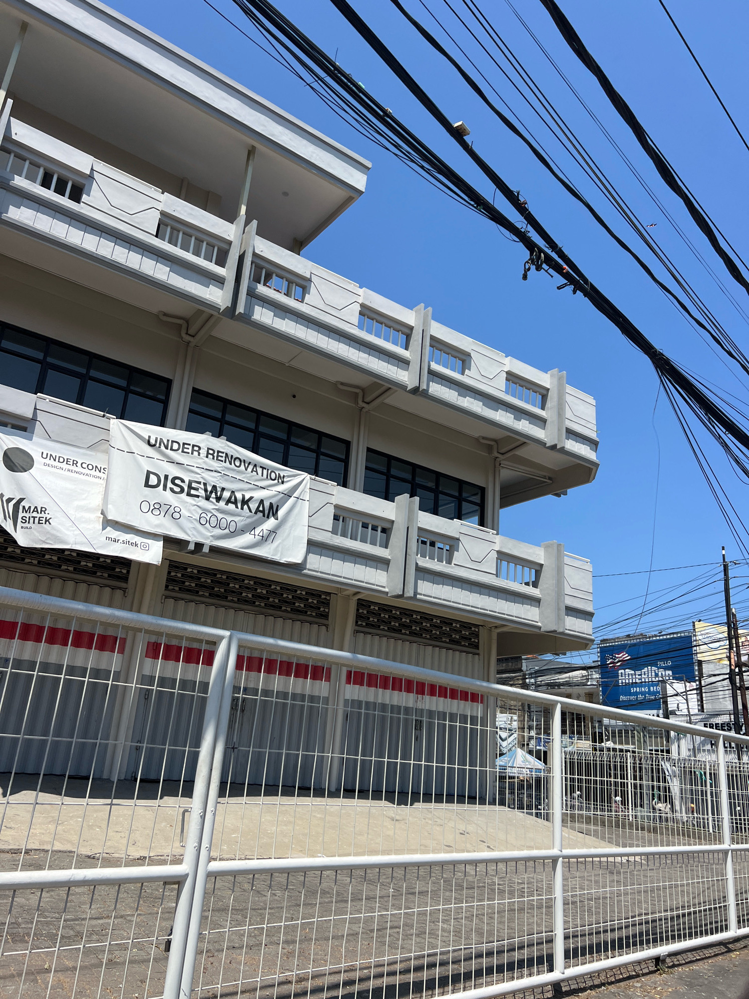 A daytime, eye-level shot captures a multi-story commercial building under renovation in Denpasar, Indonesia. A white banner across the building's facade reads "UNDER RENOVATION DISEWAKAN," with a phone number below. Another banner on the left side shows "MAR.SITEK BUILD" and "mar.sitek". A white metal fence runs along the foreground, obscuring the building's lower level which features closed roller shutters with red and white striped accents. Above the shutters are rows of windows. The sky is clear blue with a network of power lines crisscrossing the frame. In the background, a billboard advertising "American Spring Bed" is partially visible. The scene appears quiet and still, with no people actively engaged in activity within the frame.
