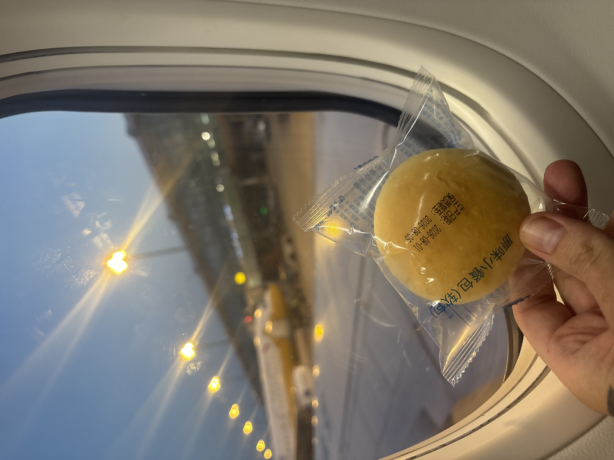 A first-person view from an airplane window shows a hand holding a packaged bread roll with Asian characters and a date "2026-08-01" printed on it. Outside the window, an airplane with yellow and white livery is parked at an airport gate. The scene is illuminated by airport lights, and the sky is a dusky blue. The location is identified as Xinwu District, China.