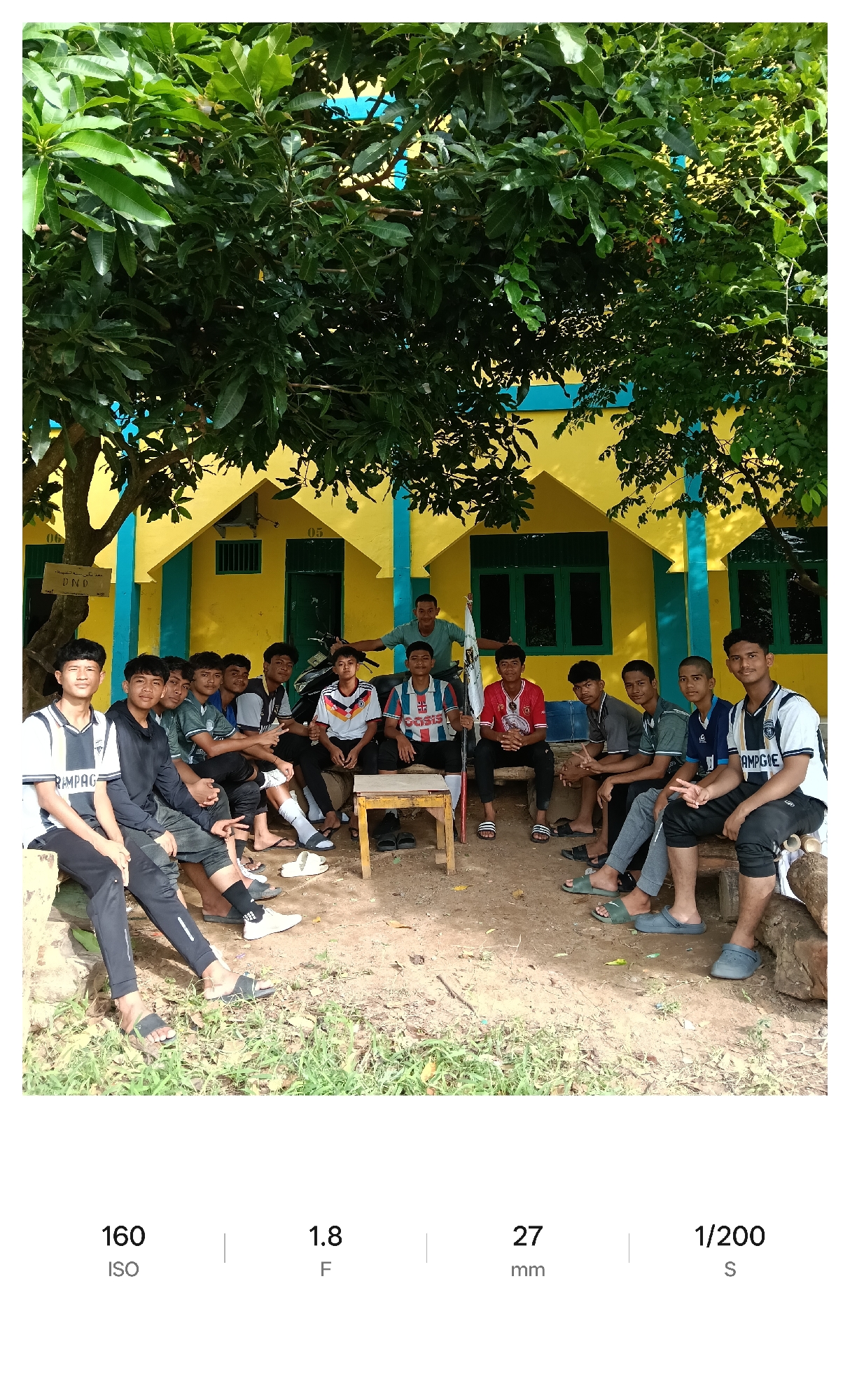 A group of approximately eleven young men, some wearing t-shirts with various logos, are gathered in front of a yellow building with blue trim and dark green windows. Some are seated on wooden benches and a small wooden table is in the center of the group. A man stands behind the seated men, holding a white flag with black writing. The scene appears to be outdoors, with a large tree providing shade. The setting is rural, with a dirt and grass ground. The sign above the door says: "هذا مكن سنة السلفية DND." The numbers 05 and 06 can be seen on the facade of the building. Based on the DND sign, the location may be near or in Lhokseumawe, Indonesia.