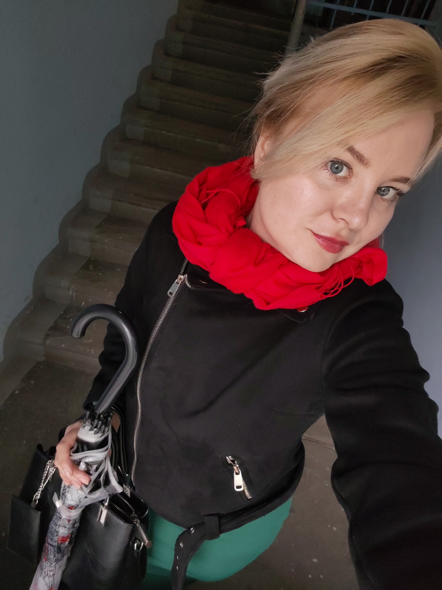 A woman is captured in an upward-angle selfie, standing on a staircase. The location is identified as Izhevsk, Russia. She has fair skin, blonde hair styled to the side, and blue eyes. She is wearing a black jacket and a bright red scarf around her neck. An umbrella is clutched in her hand, along with a black purse. The staircase is concrete and the walls are painted a neutral gray.