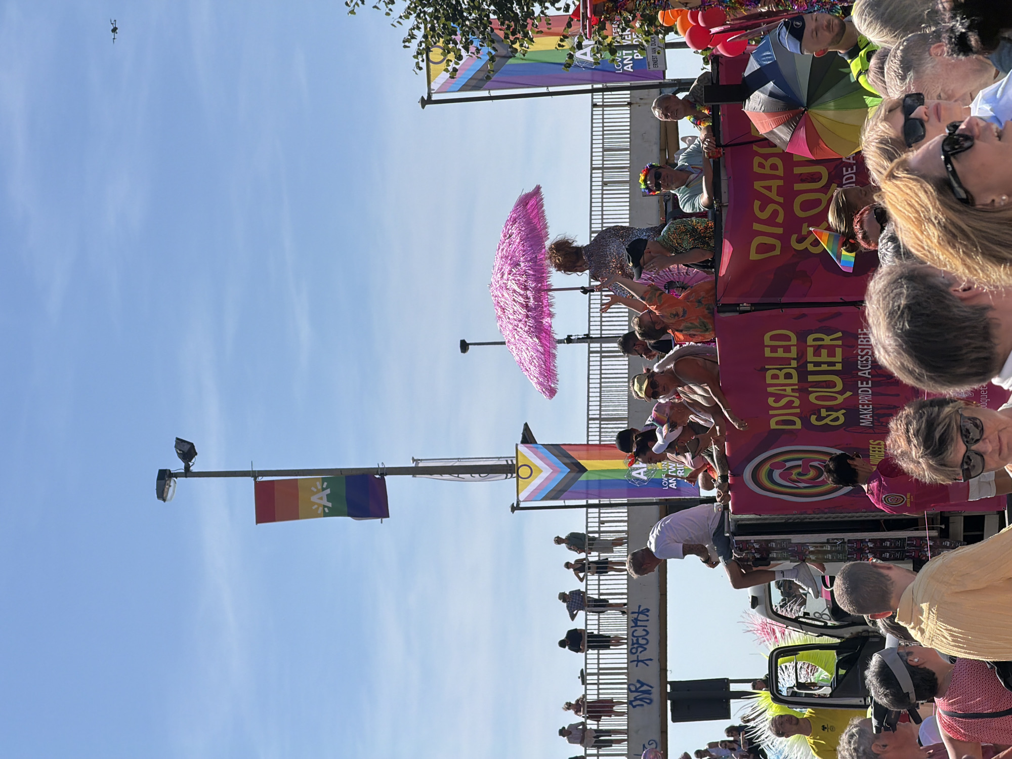 In Antwerp, Belgium, a vibrant Pride parade unfolds under a clear blue sky. A truck serves as a mobile stage, adorned with a large pink banner proclaiming "DISABLED & QUEER. MAKE PRIDE ACCESSIBLE." On the truck, individuals engage in the festivities; one person, dressed in a shimmering silver outfit, holds a pink, feathery umbrella, while others wave and interact with the crowd. A rainbow-colored umbrella is also visible. Above, a lamppost displays a rainbow flag with an "A" logo, symbolizing Antwerp's Pride event. Further in the background, spectators line a bridge, observing the parade. The scene captures the celebratory and inclusive spirit of the event.