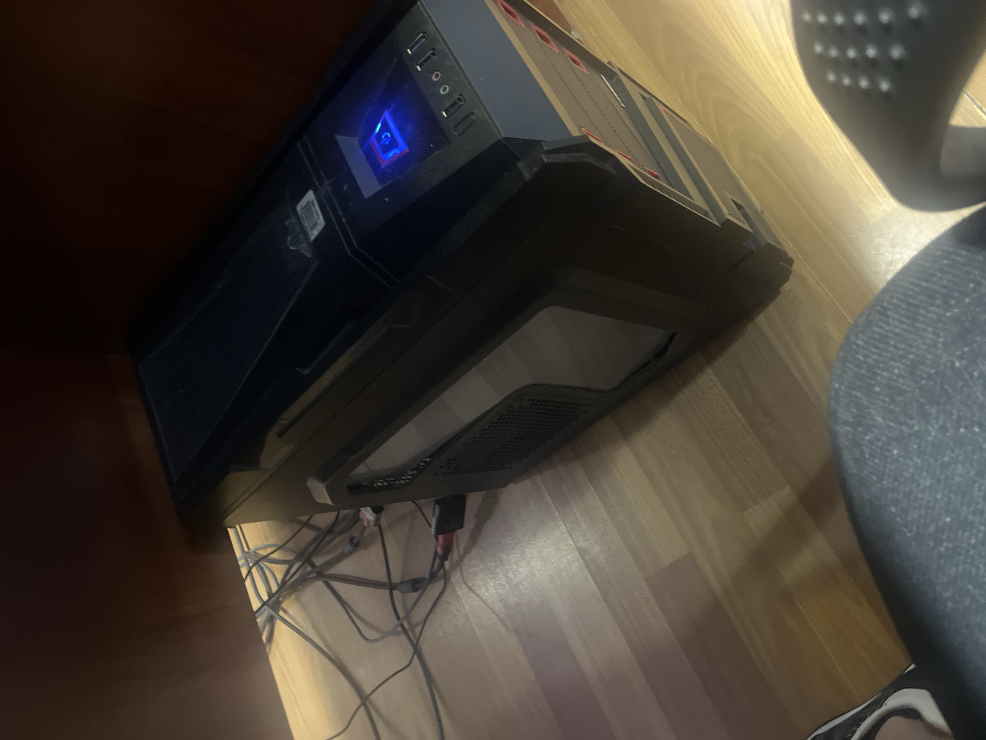 A black computer tower is positioned on a light-colored wooden floor beneath a dark brown wooden desk. Various cables are connected to the computer and are draped across the floor. The computer has a blue illuminated power button on its front panel. A portion of a person's leg, clad in blue denim jeans, and a foot wearing a black and white sneaker are visible in the foreground, suggesting they are seated or standing near the computer. The scene appears to be indoors, possibly in a home or office setting. There is no information in the image to determine the location in the city of Masis, Armenia.