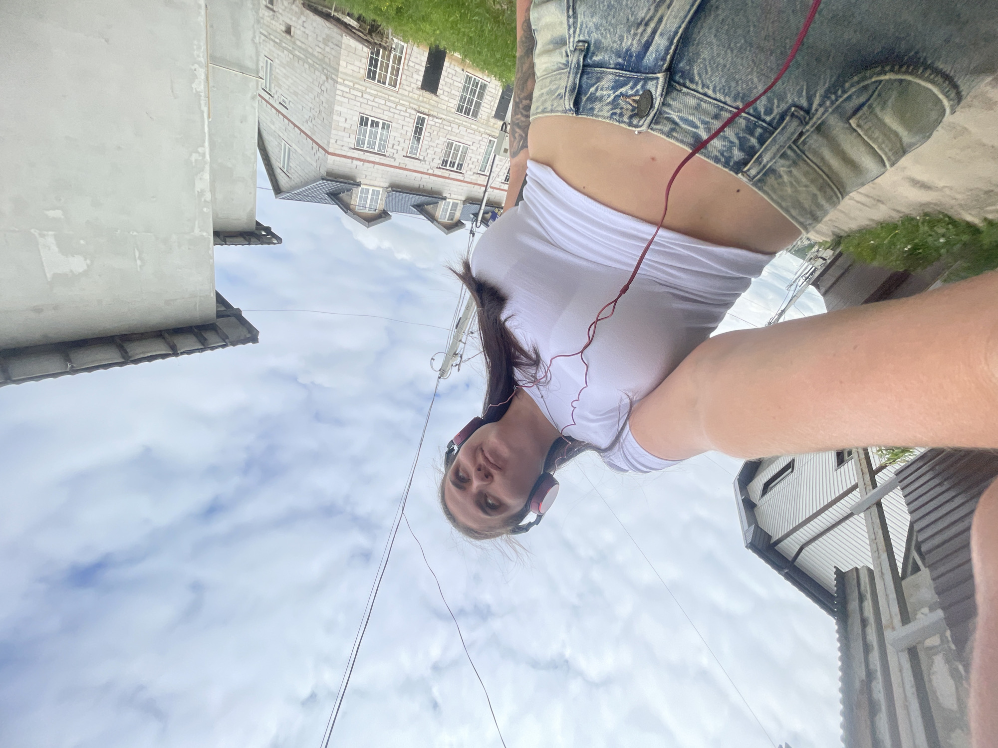 The image captures a low-angle outdoor selfie featuring a young woman against a backdrop of residential buildings under an overcast sky in Molokovo, Russia.
The primary subject is a young woman, visible from her mid-torso up, with long dark hair. She is wearing a white, form-fitting crop top and light-wash denim shorts or jeans. Large headphones with prominent red wires are on her head, extending downwards out of frame. A tattoo is visible on her left arm. She maintains a neutral expression, looking directly towards the camera, suggesting she is taking the photo herself.
The background consists of several buildings. To the left, a multi-story structure built from light-colored masonry blocks, possibly under construction or renovation, features multiple white-framed windows and a dark roof. Some green grass is visible behind or on top of this building. To the right, another residential-style building with light siding and a dark roof is partially visible, along with what appears to be a corrugated metal fence or structure in the foreground. The sky above is largely covered with scattered white clouds, indicating a daytime setting.
The image suggests the woman is possibly listening to music while posing. The perspective is dynamic, looking upwards from a low point. No visible text is present in the image.