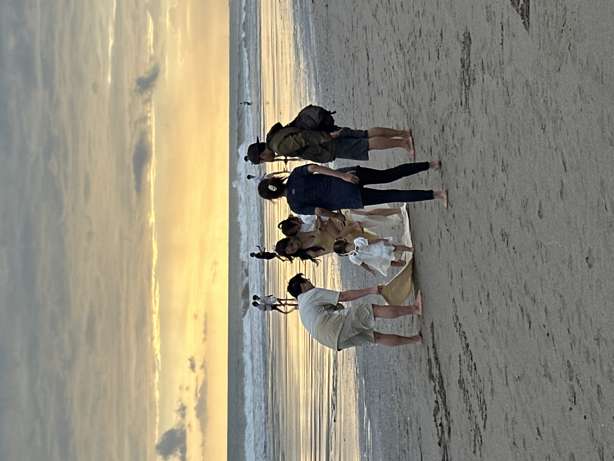 In the city of North Kuta, Indonesia, at sunset, a group of people is gathered on a sandy beach. The sky is filled with warm, golden hues as the sun sets over the ocean, casting reflections on the wet sand.
In the foreground, a man in a light-colored shirt and shorts is kneeling and holding a tan mat. Beside him, a woman in a yellow dress is holding a baby. A young girl in a white dress stands near the mat, looking at the man. To the right, a woman in a dark top and leggings stands facing away from the camera, and a man wearing a dark hat, olive-green jacket, and shorts stands next to her, carrying a backpack.
Further back, several other people are visible on the beach and in the shallow water. Some are walking, and others appear to be playing or swimming in the ocean, where waves are breaking. The environment is a typical beach scene with sand, the ocean, and a colorful sunset sky.