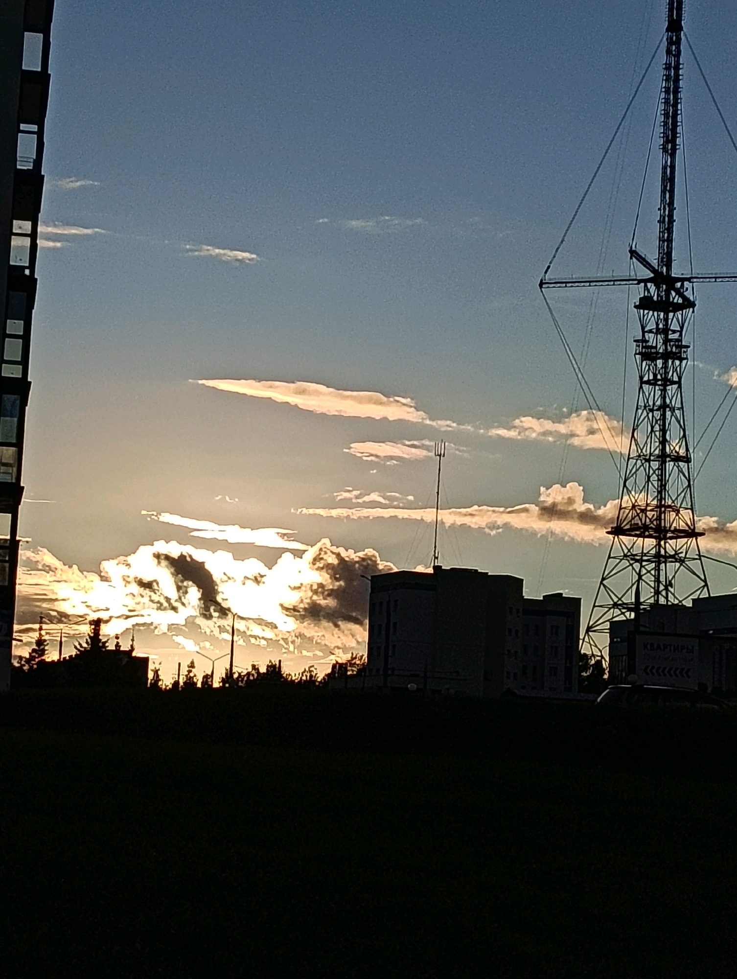 The image depicts a silhouetted cityscape against a dusky sky with dramatic clouds illuminated by the setting sun. A tall radio tower dominates the right side of the frame, with supporting guy wires stretching across the sky. To the left, a portion of a modern building with numerous windows is visible. In the middle ground, several other buildings are silhouetted, including what appears to be a block of apartments. The foreground is a dark, featureless expanse, likely grass or a field. The overall impression is one of an urban environment at dusk, with the radio tower and buildings forming a stark contrast against the illuminated sky. While the image is a general cityscape, the radio tower is likely the Hrodna Radio and Television Broadcasting Tower, a prominent landmark in Hrodna, Belarus.