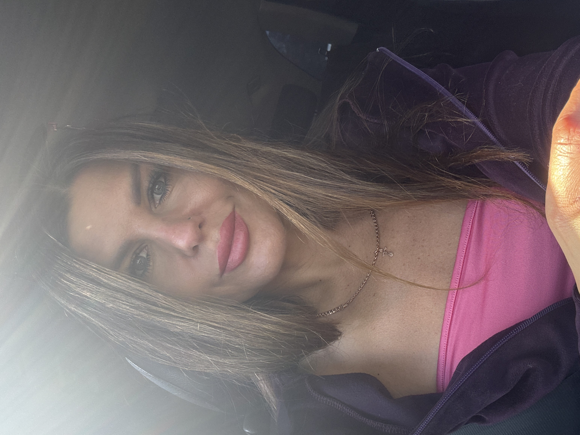 The image captures an adult female from the chest up, positioned inside what appears to be a vehicle. She is looking directly into the camera with a subtle, closed-lip smile. Her hair is long, straight, and brown with lighter highlights, draped over her shoulders. She wears a bright pink strapless top, partially visible beneath an open, dark purple or plum-colored velour hoodie with a zipper. A thin, light-colored metallic chain necklace with a small cross pendant hangs around her neck. A small metallic stud is visible in her left nostril. Strong direct sunlight from the upper left creates a lens flare and illuminates the left side of her face, hair, and a hand in the lower right corner, while the right side of the image remains darker. The interior of the vehicle, characterized by dark upholstery, serves as the background. There are no discernible features within the image to identify the specific geographical location as the city of Chaikovsky, Russia, or any other specific region.