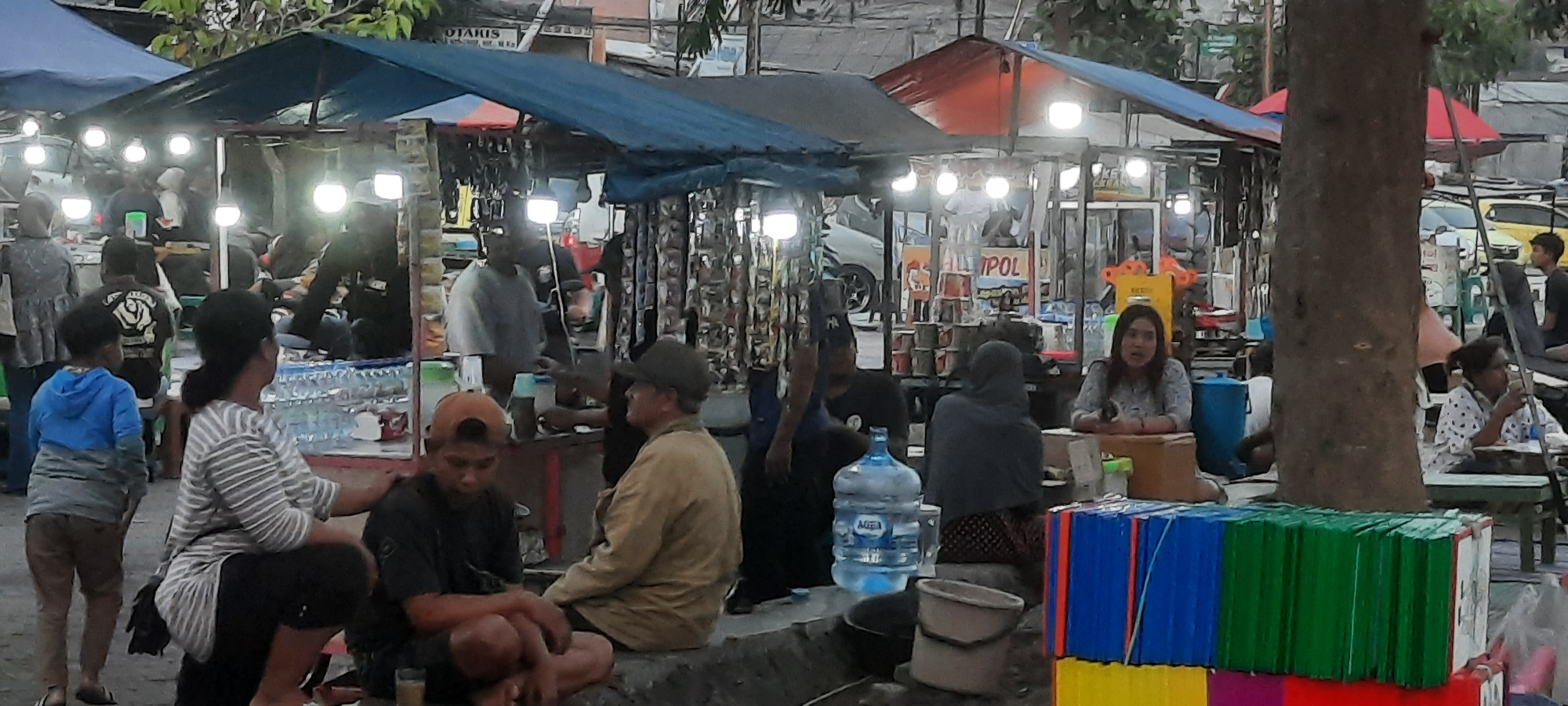 The image depicts a street-side market scene in Kanigoro, Indonesia, likely in the evening given the artificial lighting. Multiple stalls are set up, covered by blue and red tarpaulins. People are seen browsing and interacting at the stalls.
In the foreground, a woman in a striped shirt and dark pants is squatting, her left hand resting on the shoulder of a person in front of her who is facing away from the camera, wearing a blue hooded sweatshirt and brown pants. Another person, wearing a dark t-shirt and an orange baseball cap, is sitting cross-legged in the middle-left of the frame, looking down. Beside them, an individual in a tan jacket and a hat is seated, facing right and looking away.
Further into the scene, several more individuals are visible. One person, wearing a gray shirt, stands behind a stall that displays various items hanging on the left. Another stall features numerous cans stacked, with a sign that appears to read "MPOL". A woman in a patterned shirt is seated behind a counter to the right, looking towards the left. To her right, another person is seated at a table.
A large blue water dispenser jug is present near the center of the image, next to a bucket. In the background, vehicles, including a white car and a yellow car, are parked or driving on a street. The environment suggests an outdoor market with temporary structures and a generally busy atmosphere. The ground appears to be paved or a hard surface.