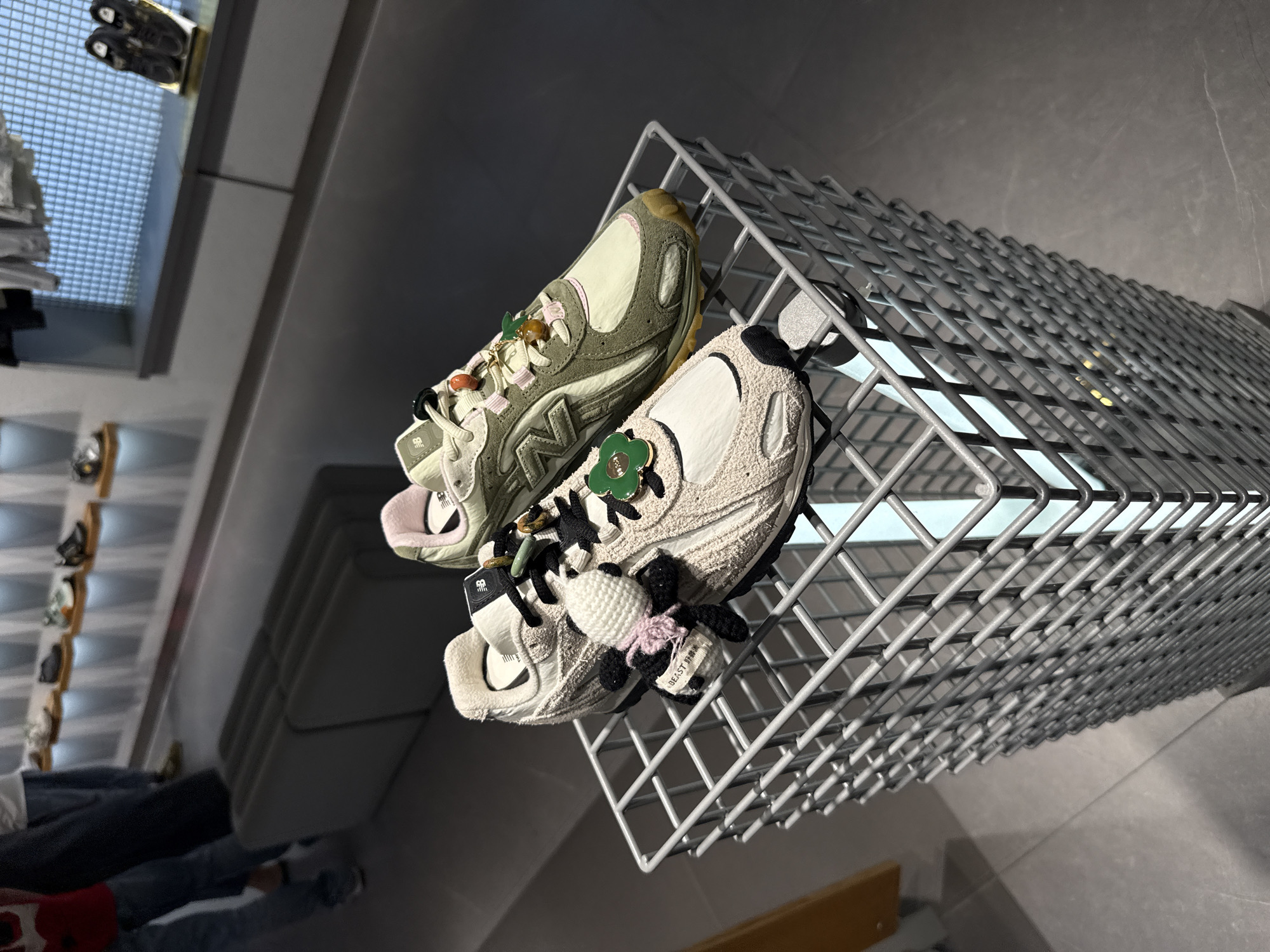 Two pairs of customized New Balance sneakers are prominently displayed on a grey metal grid shelf, likely within a retail store in Tianhe District, China. The image is taken from a low, upward-angled perspective.
The sneaker on the left features an upper constructed from olive green/khaki suede and off-white mesh, with a subtle pink lining visible around the ankle collar. Its laces are adorned with several decorative charms, including a green leaf, a brown bead, and an orange bead, alongside other small metallic elements. The "NB" logo is visible on the side of the shoe.
The sneaker on the right exhibits a cream/off-white textured upper, possibly fleece or chenille, with contrasting black details on the sole and heel. This shoe is also customized with charms on its laces: a green flower with a black center, and a distinctive white and pink crocheted animal, resembling a panda or similar creature. A small white tag attached to the crocheted charm clearly shows the English text "DEAST" and Chinese characters, which appear to be "好運" (meaning good luck). This shoe also features the "NB" branding.
In the blurred background, other shoes are visible on display racks, and the partial, out-of-focus figure of a person can be seen in the lower-left quadrant, indicating an indoor retail or showroom setting. The lighting suggests an artificial indoor environment. The image highlights the detailed and personalized elements of the footwear.