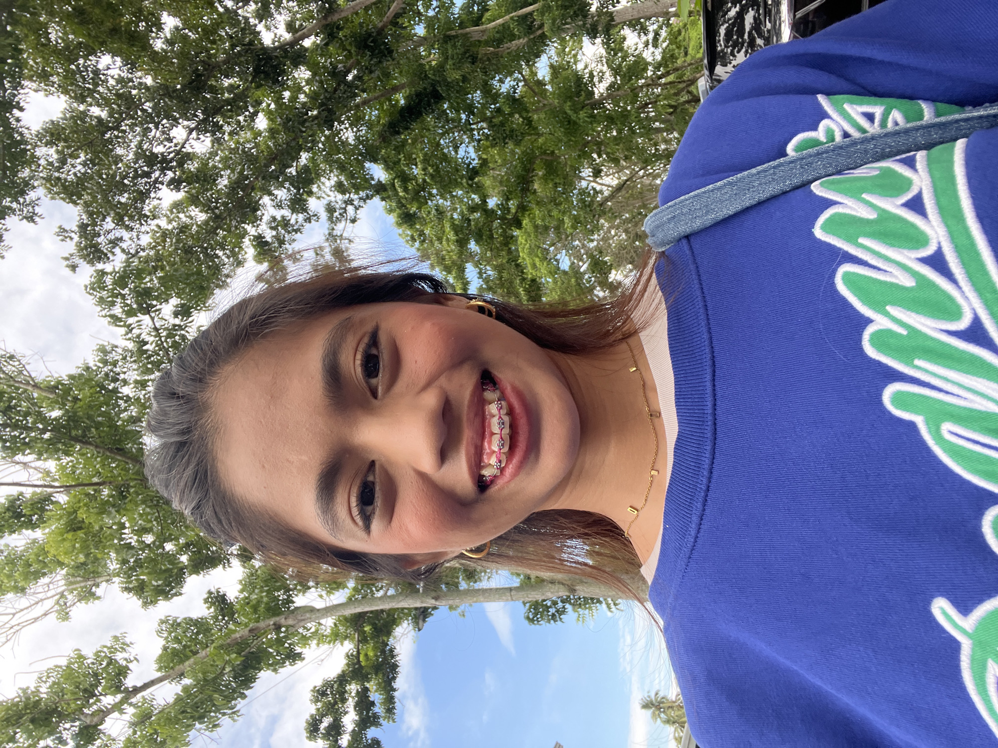 A young woman with braces and prominent blush is posing for a selfie outdoors. She is wearing a blue sweatshirt and a gold necklace. Behind her, trees and a cloudy sky are visible. The scene appears to be in Panabo City, Philippines, suggested by the presence of palm trees.