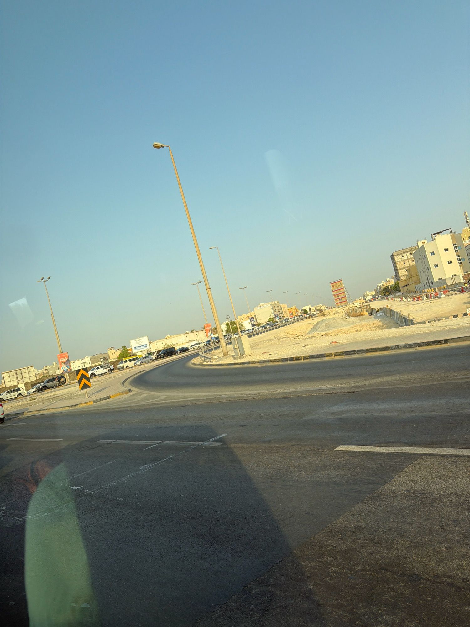 The image depicts a street scene in Riffa, Bahrain, under a clear blue sky. The foreground is dominated by a slightly tilted asphalt road with faded white lane markings and a prominent shadow cast across it. To the left, a curved road leads away, bordered by a yellow and black striped barrier and a speed bump.
Further into the scene, a construction site is visible on the right, characterized by mounds of sand, various barriers, and unfinished buildings in the background. Several vehicles, including cars and vans, are present on the road and parked along the sides. Streetlights stand tall along the road. Advertisements and traffic signs are also scattered throughout the area, including a large billboard with an image of a person. The overall impression is one of an active urban environment with ongoing development.