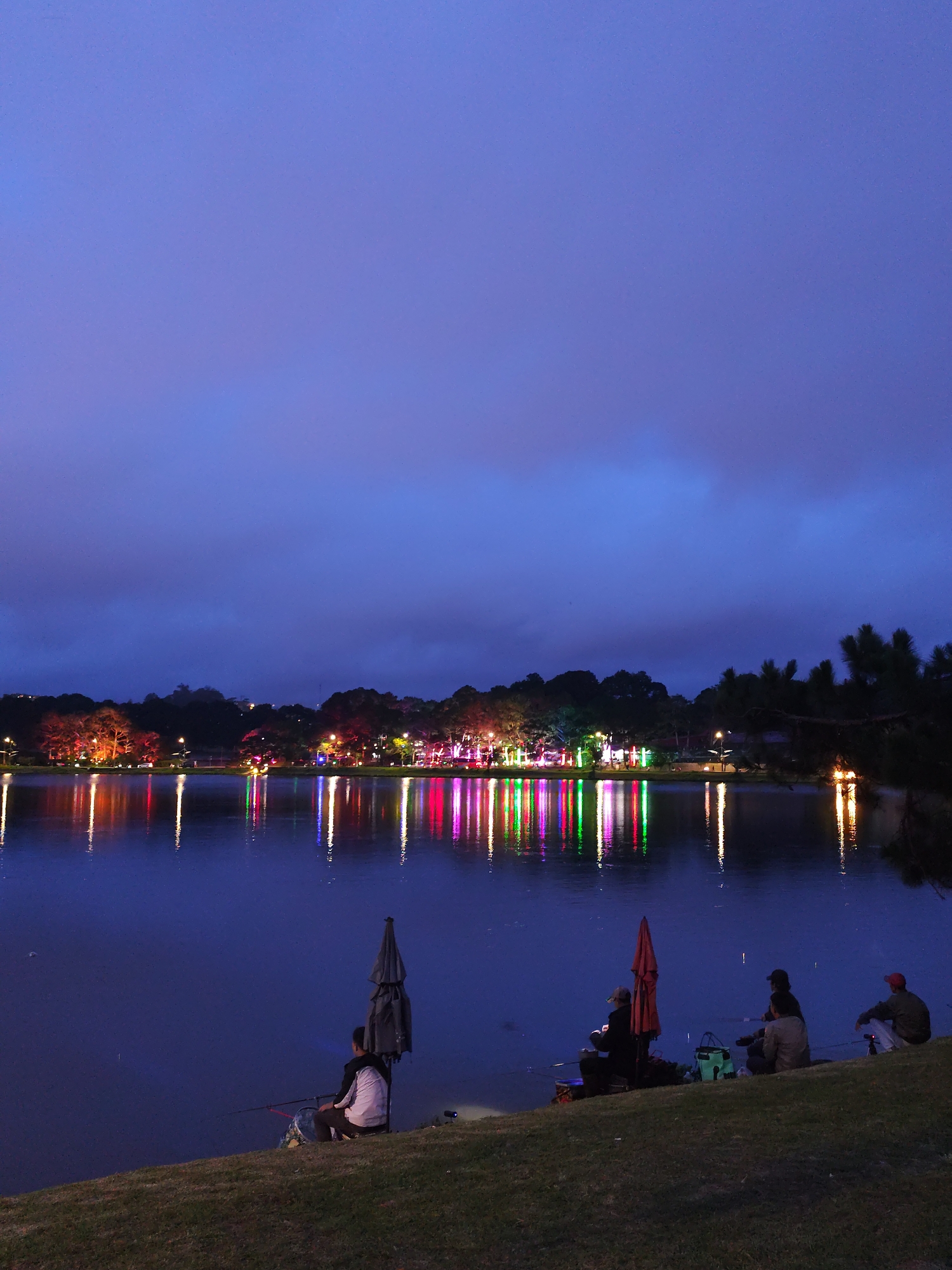 At night in Da Lat, Vietnam, five people fish on the grassy bank of a body of water, likely Xuan Huong Lake. Each person sits, with fishing rods extended towards the water. Two have dark umbrellas beside them, one red and one dark grey. Across the water, a row of lights is reflected in the dark surface. The sky is deep blue. The foreground is grassy bank and the water. The background is the sky and the tree-lined area behind the lake.