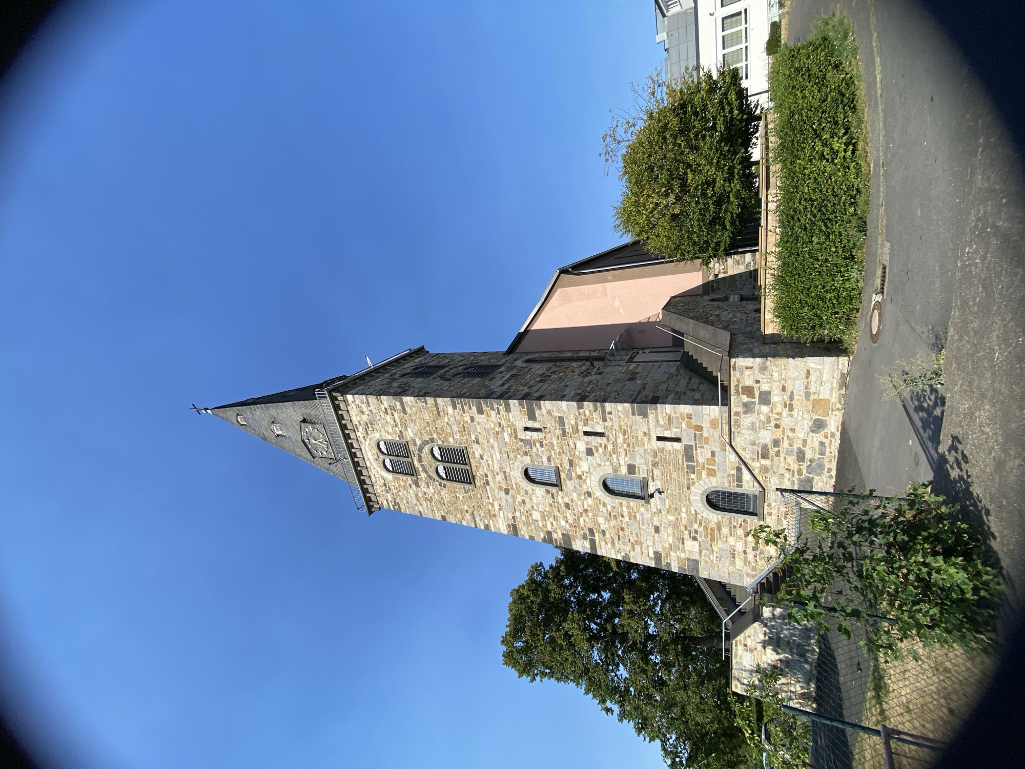 An eye-level, wide-angle shot captures the St. Nikolaus Church in Altenkirchen, Germany, under a clear blue sky. The church's prominent stone tower, topped with a dark steeple, dominates the frame. The tower features several arched windows and a clock face near the top. To the right of the tower, a section of the church building with a light pink facade is visible, with an external staircase leading up. Lush green trees and a neatly trimmed hedge surround the church. In the foreground, a chain-link fence with some overgrown green foliage runs across the lower left portion of the image. Paved ground and a manhole cover are also present in the foreground. The scene appears to be a sunny day, with shadows cast on the ground, indicating the time of day is likely morning or afternoon. There are no people visible in the image.