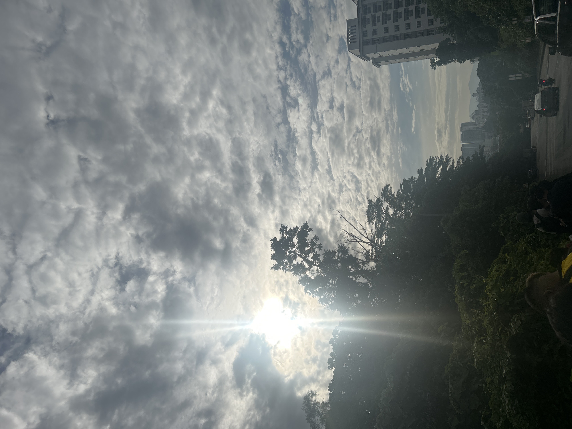 The image shows a view looking up at a cloudy sky with the sun shining through the clouds. The sun's glare creates bright streaks across the frame. In the middle ground, there are silhouetted trees. In the background, a tall building and other city structures are partially visible. There is a road on the right side with cars. The location is most likely in Subang Jaya City Council, Malaysia.