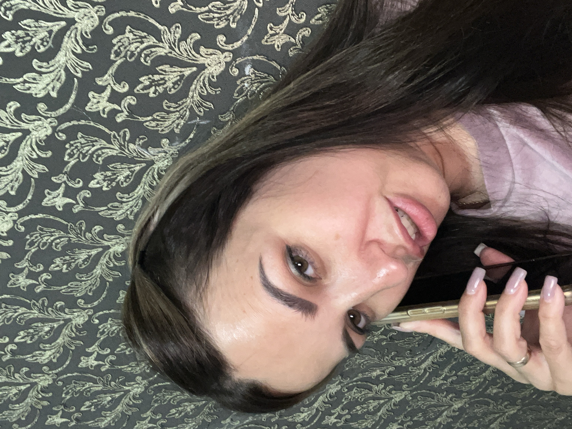 A medium shot photo shows a woman with dark hair holding a gold-colored smartphone to her left ear. She is wearing a light purple t-shirt and a gold ring on her left hand. The woman is looking at the camera with her mouth slightly open. Her makeup includes eyeliner and lipstick. The background is a dark gray wallpaper with a gold floral pattern.
